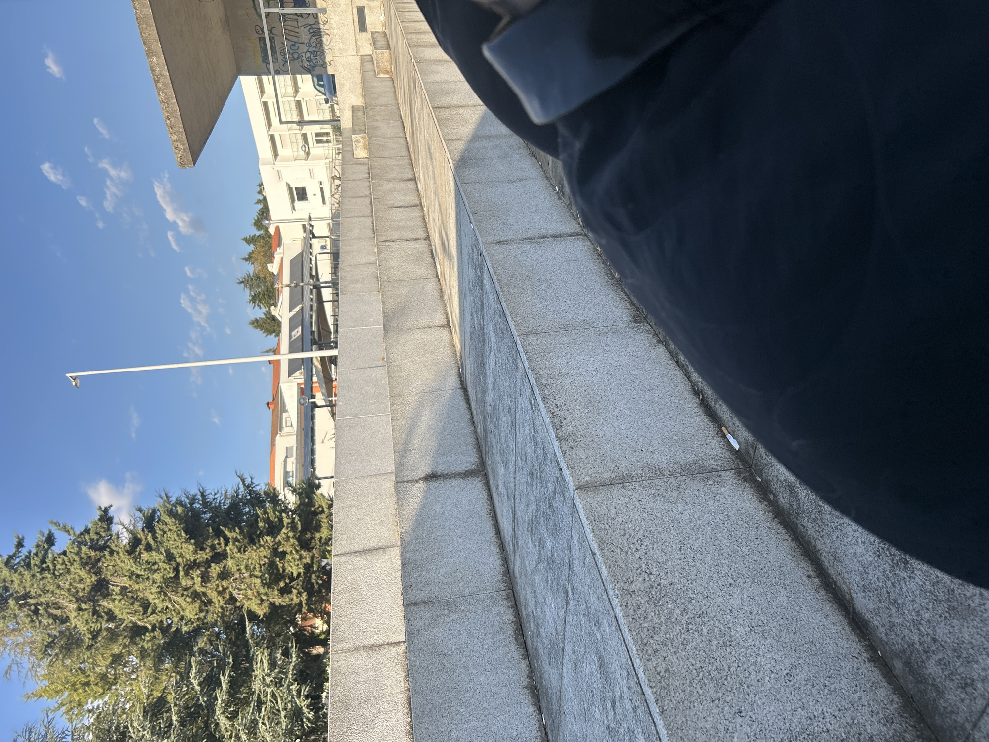 The image shows a sidewalk or stairs made of gray stone in Vila Real, Portugal. A portion of a person wearing dark clothing is visible on the right side of the frame, possibly obscuring a full view of the scene. The perspective is tilted.
In the background, there are residential buildings, trees, and a clear blue sky with scattered clouds. A light pole is visible. There's also graffiti on the building in the background.