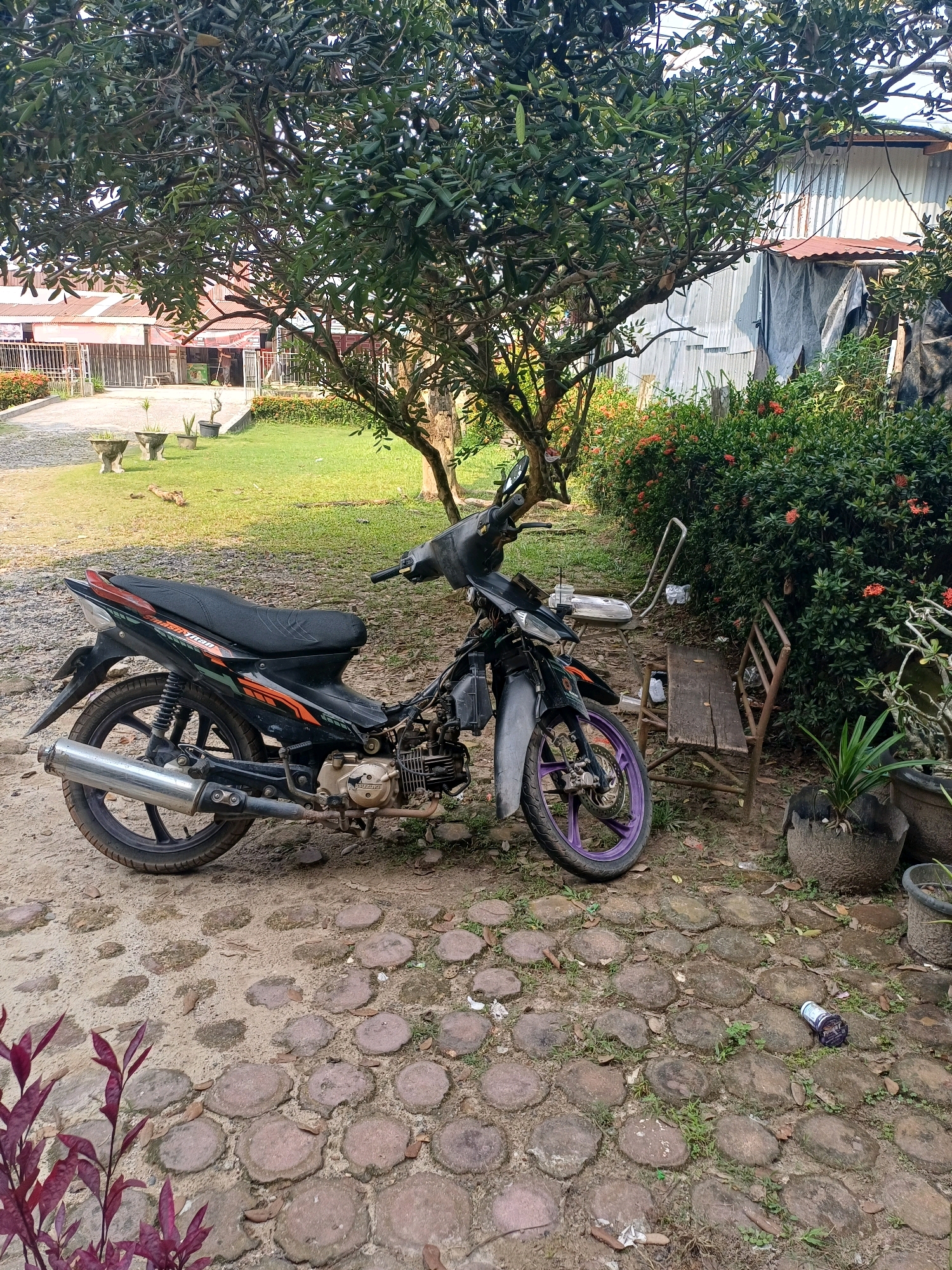 A black motorcycle is parked on a dirt and stone pathway next to a lush green hedge with red flowers. Behind the hedge, a weathered wooden bench is visible, with a metal folding chair nearby. Further in the background, a building with a corrugated metal roof stands under a large tree. The scene is set in an outdoor environment, likely a residential area, in Dumai City, Indonesia. The time of day appears to be daytime, with bright sunlight casting shadows. No people are present in the image.