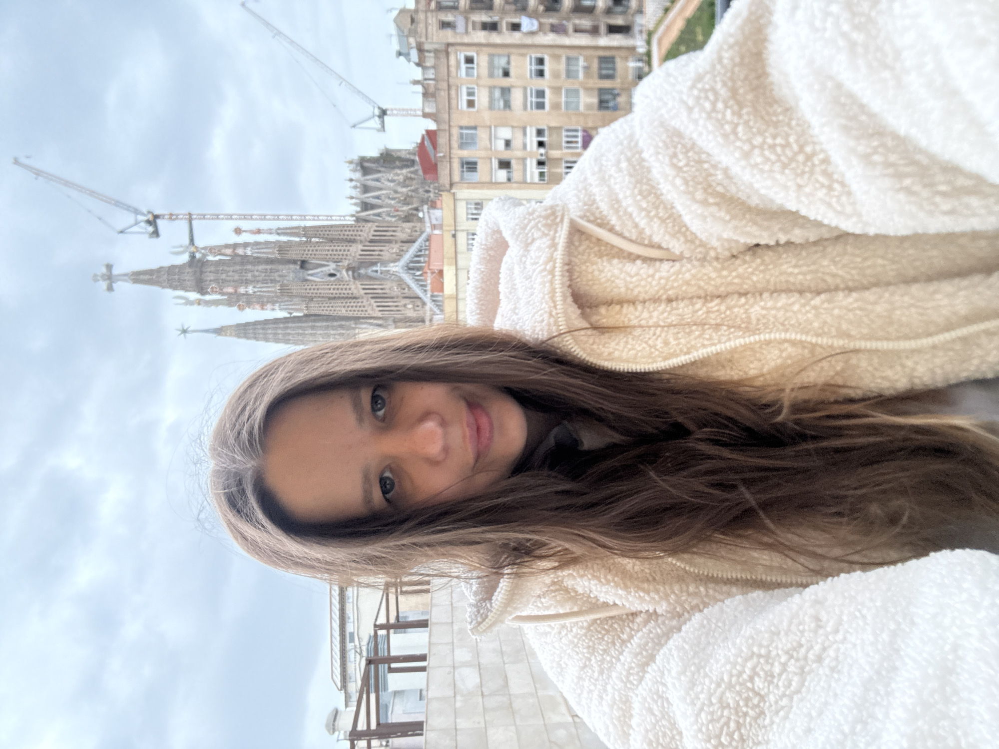 A woman takes a selfie in front of the Sagrada Familia in Barcelona, Spain. The unfinished basilica is in the background, along with construction cranes and nearby buildings. The sky is overcast. The woman is wearing a white, furry jacket.