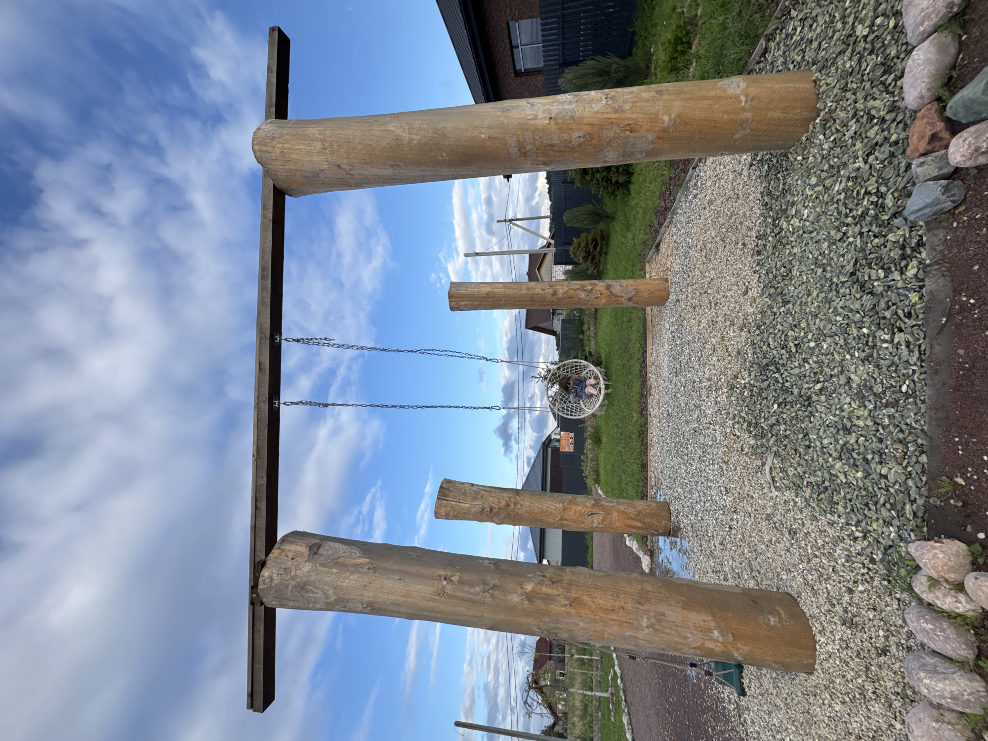 An outdoor, eye-level, wide-angle photograph captures a rustic swing set constructed from large, natural wooden logs in Timmolovo, Russia. The swing set features a horizontal wooden beam supported by two large, upright log posts. Two chains descend from the beam, holding a round, white woven swing seat suspended in the center. A young child, wearing a blue dress, is seated in the swing. The ground beneath the swing set is covered in a mixture of light and dark grey gravel. To the left of the swing set, another smaller, rough-hewn log stands vertically. In the background, a neatly manicured lawn transitions into a dark wooden fence. Beyond the fence, a brick house with a dark roof is visible, along with other residential buildings and trees under a blue sky with scattered white clouds. The overall impression is one of a natural and slightly unfinished playground setting.