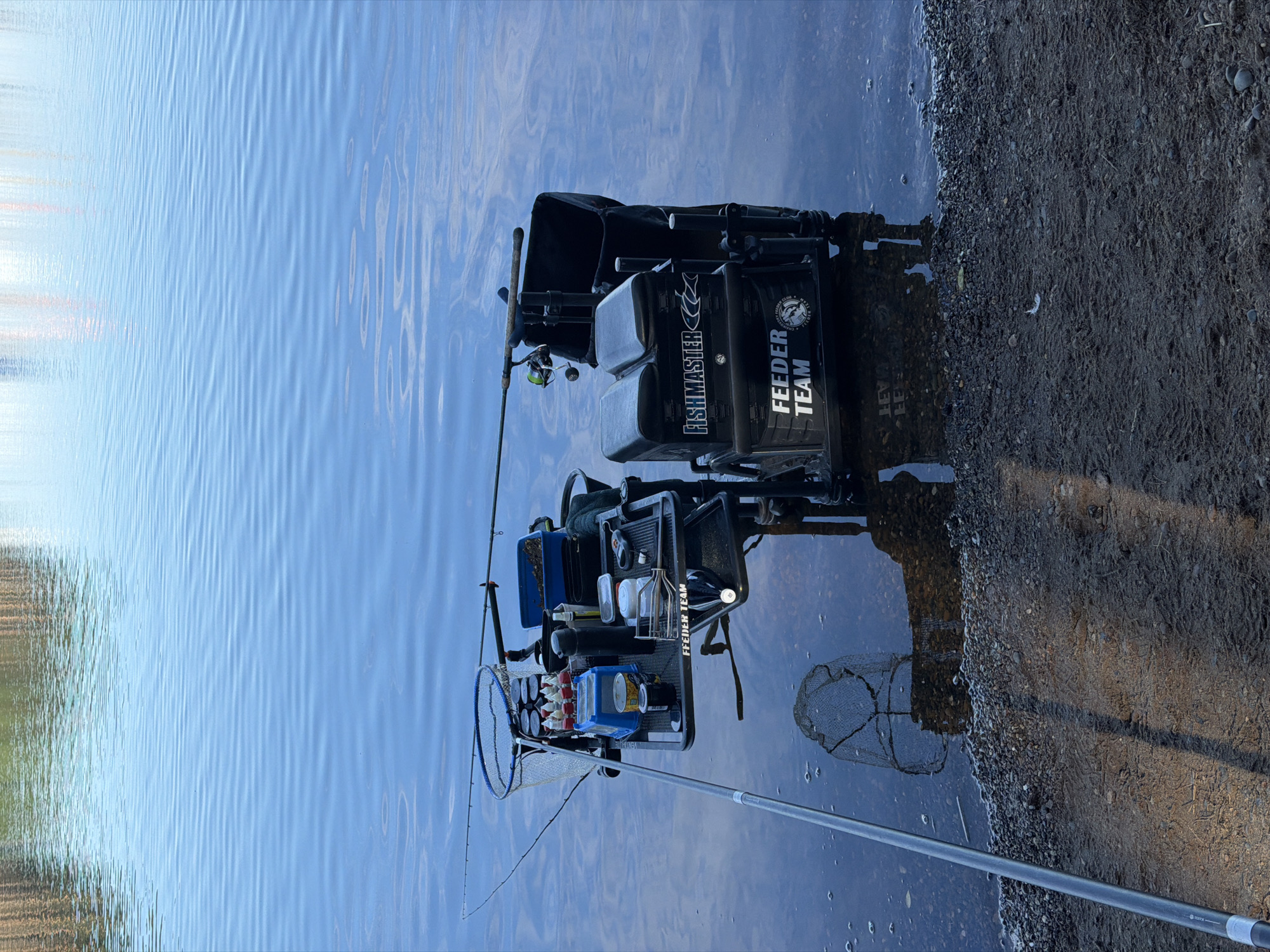 An outdoor, eye-level, medium shot shows fishing equipment set up by the water's edge in Chaikovsky, Russia. The scene is dominated by a dark-colored fishing seat box with "FISHMASTER" and "FEEDER TEAM" logos. Various fishing accessories, including containers with bait, a fishing net, a thermos, and fishing rods, are arranged on a platform attached to the seat box. One fishing rod extends from the right side of the frame towards the water. Another long fishing rod is angled from the left side of the frame, its tip in the water. The water is calm with slight ripples. The shoreline is composed of gravel and wet sand, with reflections of the equipment visible in the water. The overall atmosphere suggests a fishing activity in progress.