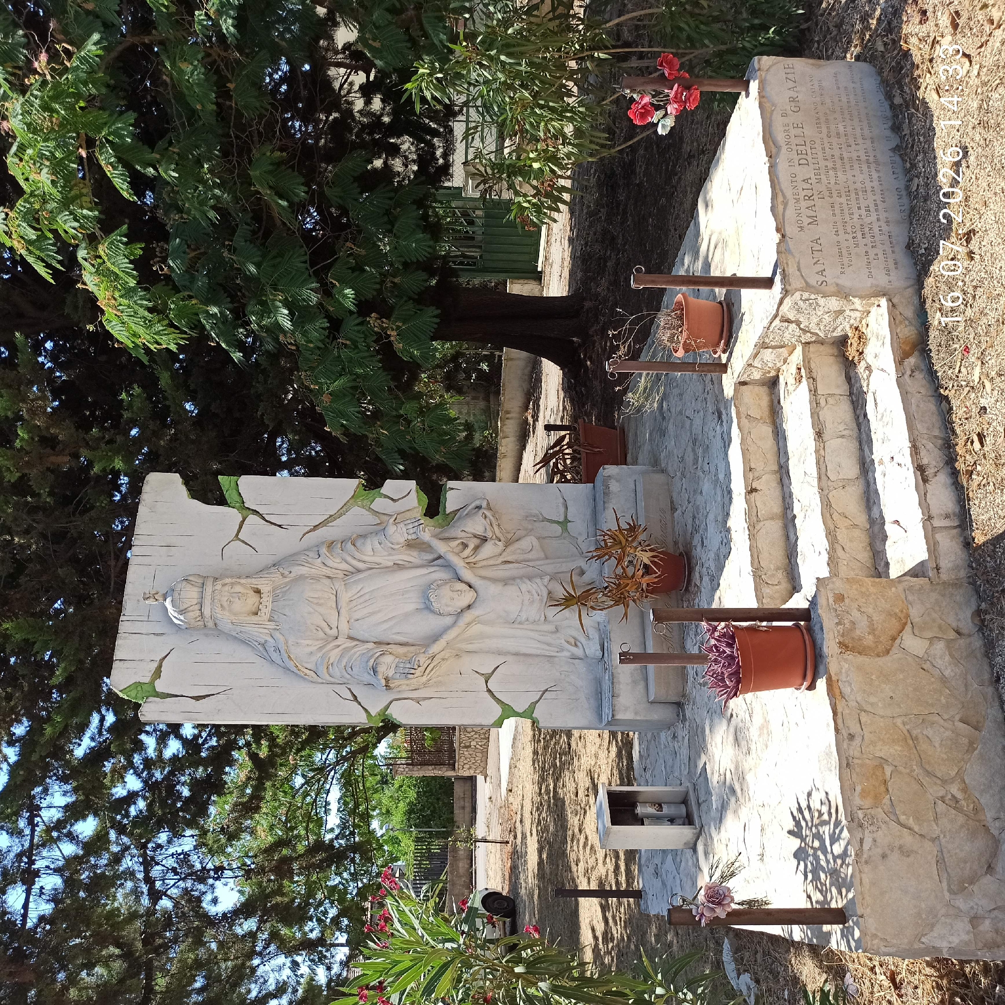 The image shows a stone monument dedicated to "Santa Maria delle Grazie" in Mellitto, Grumo Appula, Italy. The monument features a relief sculpture of a woman, presumably the Virgin Mary, wearing a crown and holding the hand of a child. The sculpture is set against a white, cracked background that appears to be made of plaster or concrete. The monument is situated outdoors in a park-like setting with trees and vegetation. There are steps leading up to the monument, and several potted plants are placed around the base. An inscription on the monument states it was realized by the local committee and dedicated to all mothers and their children. The date "16.07.2026 14:33" is superimposed on the image, suggesting it was taken on this date and time. In the background, a fence, a building, and a green gate are visible. The overall scene conveys a sense of reverence and remembrance.