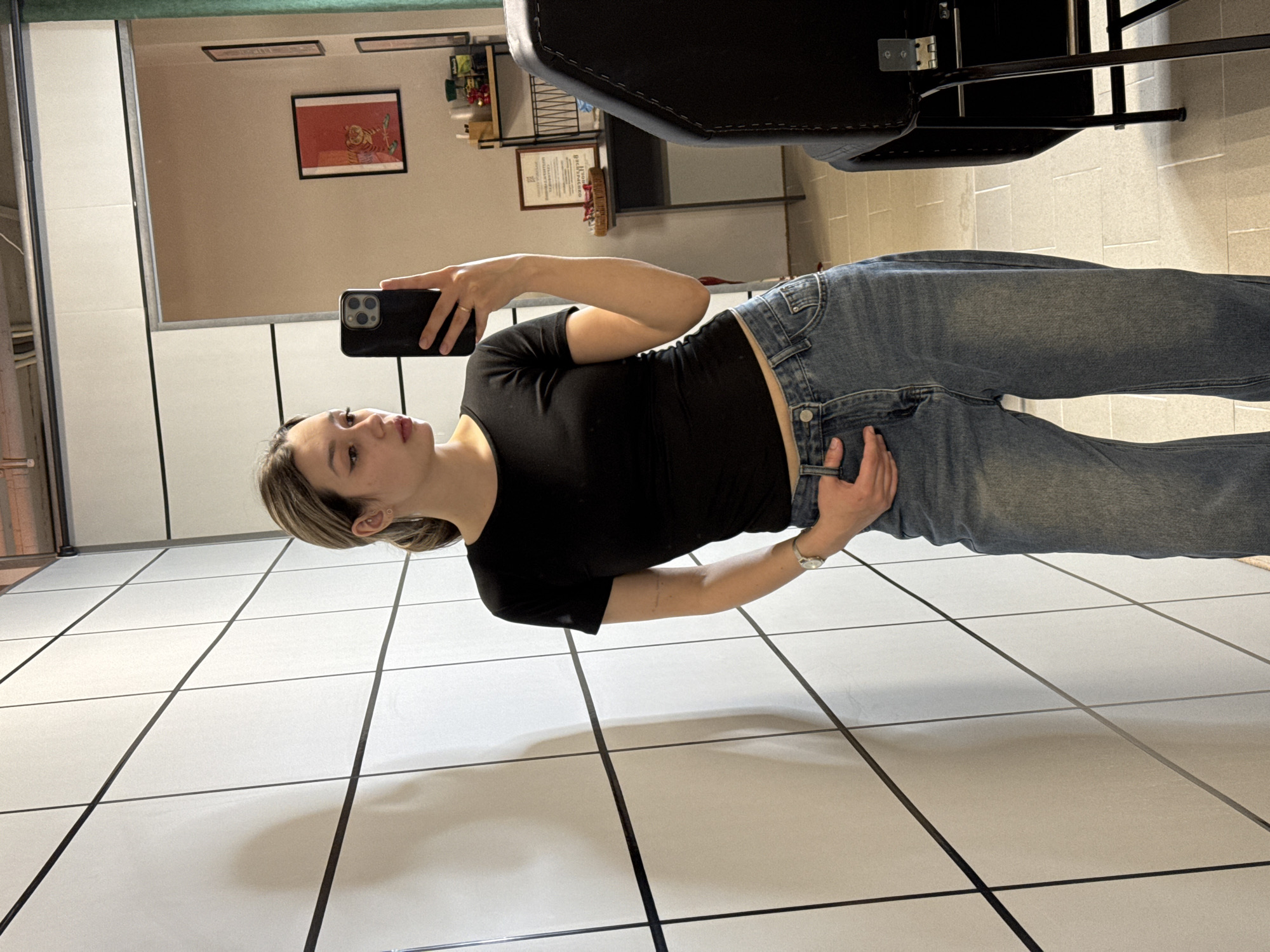 An image captures a young woman taking a mirror selfie indoors. The photograph is oriented sideways, with the woman appearing to stand horizontally.
**Subjects:** The central subject is a young woman with medium-length, light brown hair that has lighter streaks. She is wearing a fitted black short-sleeved t-shirt and blue denim jeans. A silver-toned watch is visible on her left wrist. She holds a black smartphone with a triple camera array in her left hand, positioned to capture her reflection. Her right hand is casually tucked into the waist area of her jeans.
**Scene and Setting:** The woman stands on a white tiled floor with dark grid lines, which runs vertically across the frame. The background behind her is a mirror reflecting an interior space. This reflected room contains what appears to be light-colored walls, shelves holding various items, and framed artwork. One prominent piece of art shows a tiger-like creature on a red background. Part of a dark-colored chair or furniture item is also visible in the reflection above the woman.
**Activity:** The woman is actively engaged in taking a photograph of herself using a mirror. Her gaze is directed towards her reflection and the phone.
**Notable Details:** The lighting is consistent with an indoor setting, and soft shadows are cast on the tiled floor. The woman's expression is neutral and focused. Small earrings are visible in her left ear. There is a subtle, unidentifiable tattoo on her right forearm.
**Visible Text:** No discernible text is visible on any of the objects or framed items in the reflection.