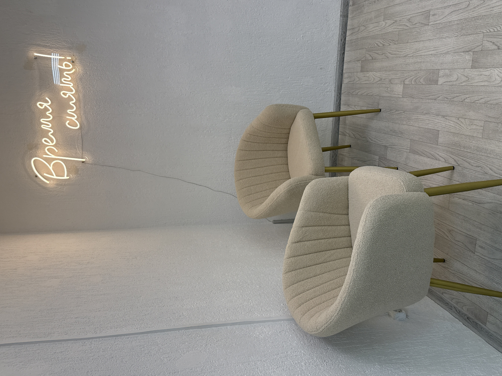 The image displays an interior space, rotated 90 degrees clockwise, giving the impression of objects defying gravity. In the foreground, two empty, modern chairs with distinctive curved backs and seats are visible. They are upholstered in a light beige, textured fabric, possibly bouclé, with horizontal stitching details. Each chair rests on thin, gold or brass-colored legs.
The setting features light-colored walls. The wall on the left side of the image (which would be the ceiling in correct orientation) is a light off-white, textured surface. A neon sign is mounted on this wall, glowing with a soft yellow-orange light. The sign displays text in Cyrillic script: "Время сиять!", which translates to "Time to shine!" A power cord trails from the sign.
The wall on the right side of the image (which would be the right wall in correct orientation) is covered with light gray, wood-like paneling, arranged horizontally, giving a clean, minimalist aesthetic. The "floor" area in the bottom part of the image is also a light, textured surface, possibly concrete or a similar finish, with a visible seam running horizontally.
No people are present, and no specific activity is taking place. The space appears clean, modern, and well-lit, suggesting a commercial or professional environment like a waiting area, salon, or office lounge in Chaikovsky, Russia. The overall atmosphere is calm and inviting.