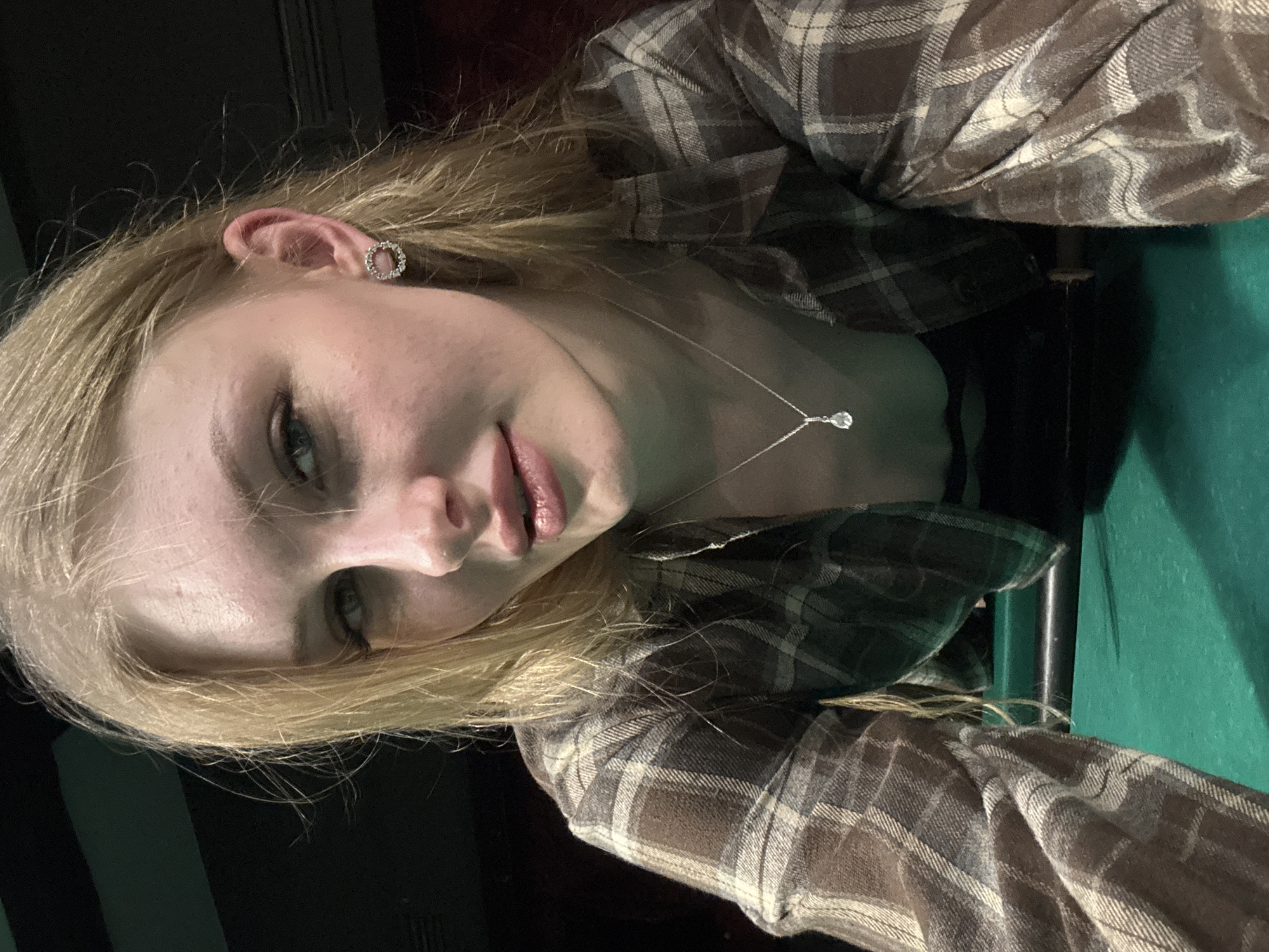 A young adult female is depicted leaning forward over a dark green felt surface, consistent with a billiards table, in an indoor setting in Perm, Russia. The subject has blonde hair, light-colored eyes, and is looking directly at the camera. She wears a brown, tan, and grey plaid long-sleeved shirt, a silver-toned necklace with a clear teardrop-shaped pendant, and a circular earring adorned with small stones in her visible right earlobe. A dark, cylindrical object is partially visible on the green surface to the right. The background is dimly lit and out of focus, with dark tones and a faint reddish-brown hue visible in sections.