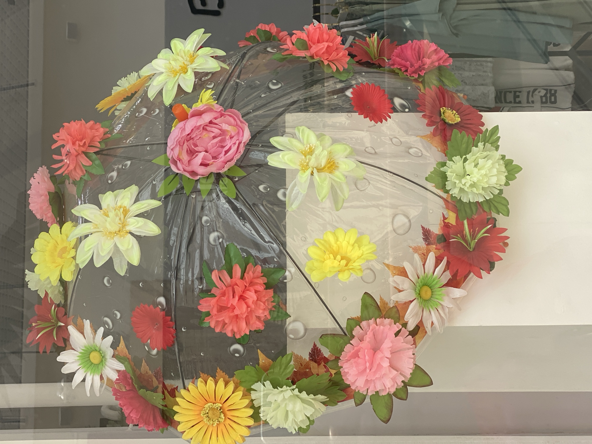 The image shows a transparent umbrella decorated with artificial flowers and autumn leaves. The umbrella is hanging, possibly in a shop window. The flowers are in various colors, including pink, yellow, red, white, and coral. The umbrella itself has a pattern of water droplets. In the background, clothing can be seen hanging, suggesting a retail environment. The context provided regarding Novokuznetsk, Russia, is external to the visual information present in the image.