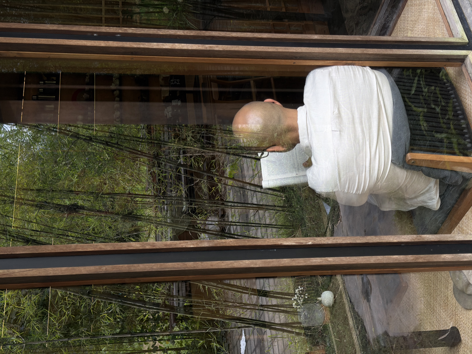 A man sits in a building, viewed through a window. The location is Sukawati, Indonesia. The man, who is bald and wearing a white shirt, is reading a book. The window reflects a dense thicket of bamboo trees and other foliage. The building's interior features wooden frames and a textured floor covering.