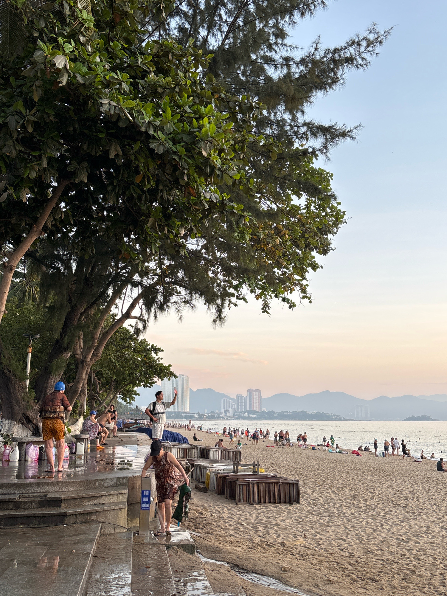 The image depicts a busy beach scene in Nha Trang, Vietnam, likely in the late afternoon, given the soft, warm light. The foreground features wet, stone steps leading down to the sandy beach. Several people are present on these steps, some appearing to be rinsing off or preparing to enter the water.
On the left, a person wearing a brown and yellow patterned vest and orange shorts, with a blue head covering, stands on the wet steps, facing away from the camera. To their right, a person in a brown patterned dress is bending down near a water spigot, holding a green garment.
Further back, a man in a white t-shirt and white shorts is standing with his arm raised, holding a mobile phone, possibly taking a picture or video. He is facing towards the ocean and the distant city skyline.
The beach itself is populated with numerous individuals, some walking along the shoreline, others sunbathing or sitting. There are various beach chairs and wooden structures scattered across the sand. In the background, the ocean stretches out towards a hazy horizon, with the silhouette of mountains and a city skyline visible. The sky is a soft gradient of pale blue and orange. Lush green trees, with broad leaves and some sparser, pine-like foliage, overhang the beach area, providing shade. The overall atmosphere is one of a popular tourist destination at the end of a day, with people enjoying the beach and the sea.