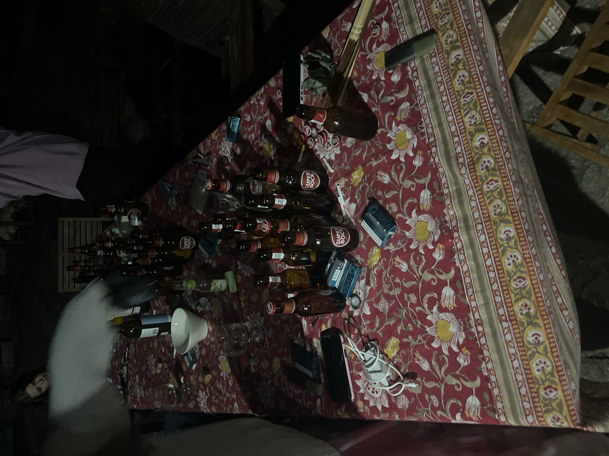 The image depicts an outdoor gathering at night, likely in the city of Felgueiras, Portugal. A long table is spread with a red floral tablecloth. Scattered across the table are numerous bottles of "Super Bock" beer, many of which appear to be empty or partially consumed. Several wine glasses and a white bowl are also visible. Other objects on the table include a mobile phone, a white charging cable and adapter, cigarette packs, and scattered bottle caps. In the background, partially obscured by a white, misty effect, a person with long brown hair is looking downwards. Another person, wearing a light purple top and dark pants, is partially visible on the right side of the frame, facing away from the camera. Wooden chairs are positioned around the table. The overall scene suggests a relaxed social gathering or party.