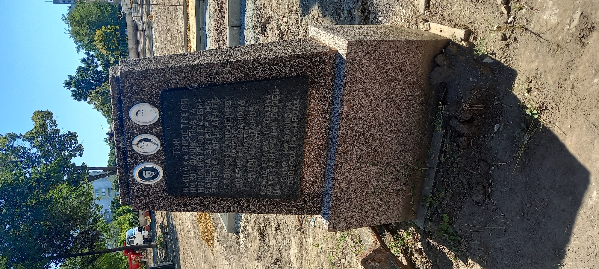 The image shows a granite memorial with three circular portraits at the top and an inscription on a black plaque below. The portraits are of young men, their faces rendered in monochrome. The inscription, in Cyrillic, appears to be a memorial to victims of fascism, with names and a date, "7-IX-1944." The monument is situated outdoors, on unpaved ground with some sparse vegetation and debris. In the background, a white truck and trees are visible, with buildings in the distance. The lighting suggests a sunny day. This memorial is located in the city of Silistra, Bulgaria, specifically in the immediate vicinity of the former prison, where victims of the communist regime were executed.