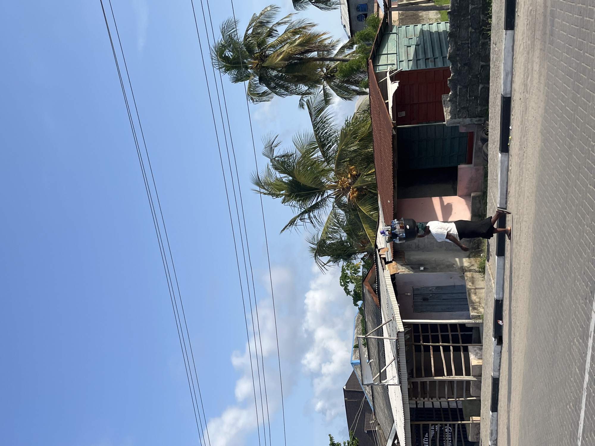 A street scene in Badagry, Nigeria, shows a person walking on a sidewalk next to a street. Overhead electrical wires run across the sky. Palm trees and various buildings are visible in the background. The person on the sidewalk is carrying something on their head. The buildings appear to be of simple construction, with a mix of materials used in their facades. The sidewalk is bordered with black and white markings.