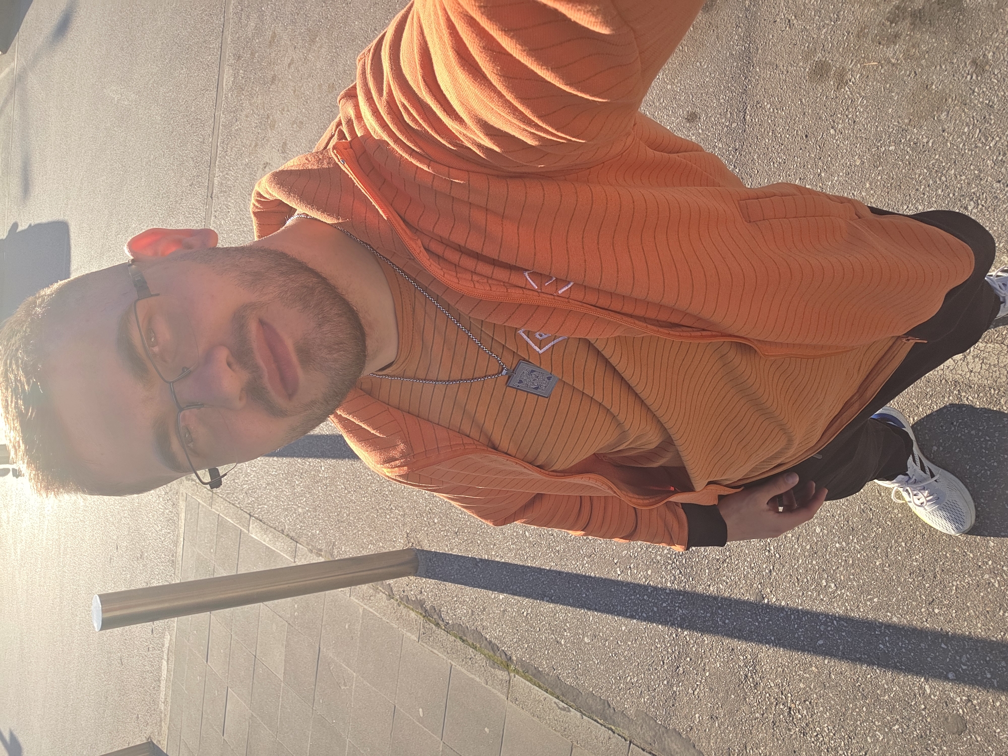 A man stands on a paved surface next to a metallic railing, likely in an urban setting. He wears glasses, an orange shirt, an orange jacket, black pants, and white sneakers. He also has a chain with a pendant around his neck. Light illuminates the left side of his face, while shadows stretch across the pavement. It is impossible to verify if the location is Plovdiv, Bulgaria.