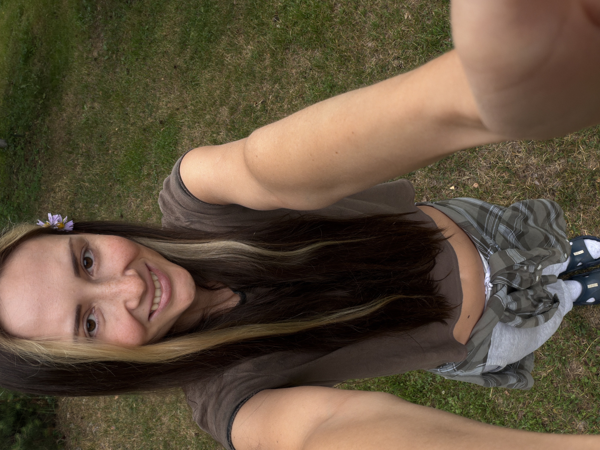 This media file is a selfie taken outdoors, likely in a park or grassy area, given the visible grass and sparse foliage. The subject is a woman with long, dark hair streaked with blonde highlights. She is smiling at the camera, and a small purple flower is tucked behind her ear. She is wearing a brown t-shirt and what appears to be a plaid shirt or skirt over gray pants. Her footwear consists of blue slide-on sandals with white socks. One of her arms is extended towards the camera, indicating the selfie was taken by herself. The lighting suggests it is daytime, and the overall mood appears relaxed and casual. The setting seems to be a natural outdoor environment, possibly in Udelnaya, Russia, as indicated by the location context. There is no visible text in the image.
