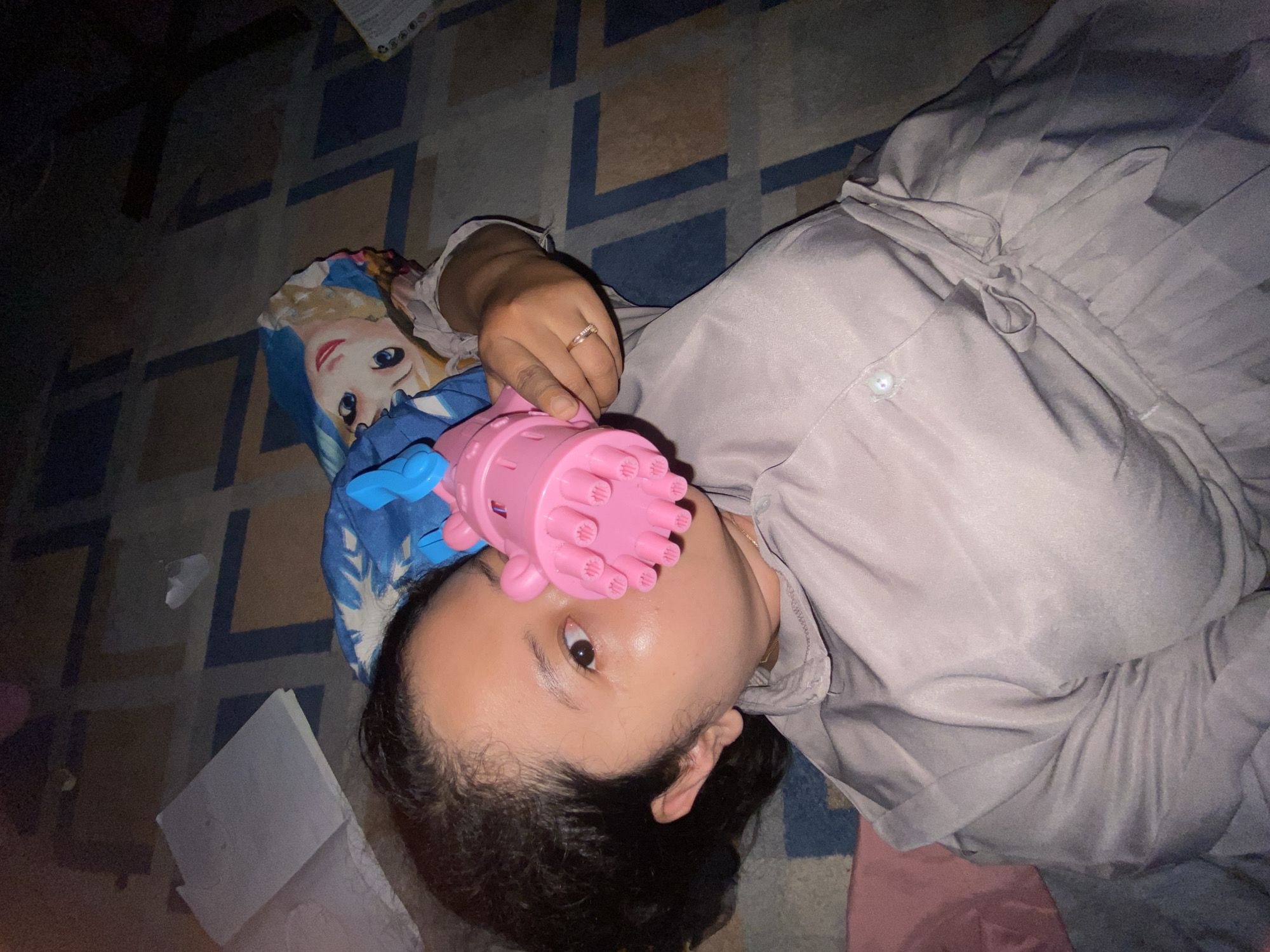 An individual is depicted lying on a patterned carpet in what appears to be an indoor setting in Gelam, Indonesia. The person, an adult with dark hair, is wearing a light purple-grey long-sleeved garment featuring buttons and a tie detail on the side. Their left hand, adorned with a ring on the ring finger, holds a pink plastic toy with multiple nubs, positioned to partially obscure the lower face and left eye. The right eye is open and directed towards the camera. A pillow featuring a cartoon character design is situated beneath the individual's head. In the background, two white books or notebooks are visible on the carpet in the upper left, while a partially obscured item with text is present in the upper right. The lighting is dim.