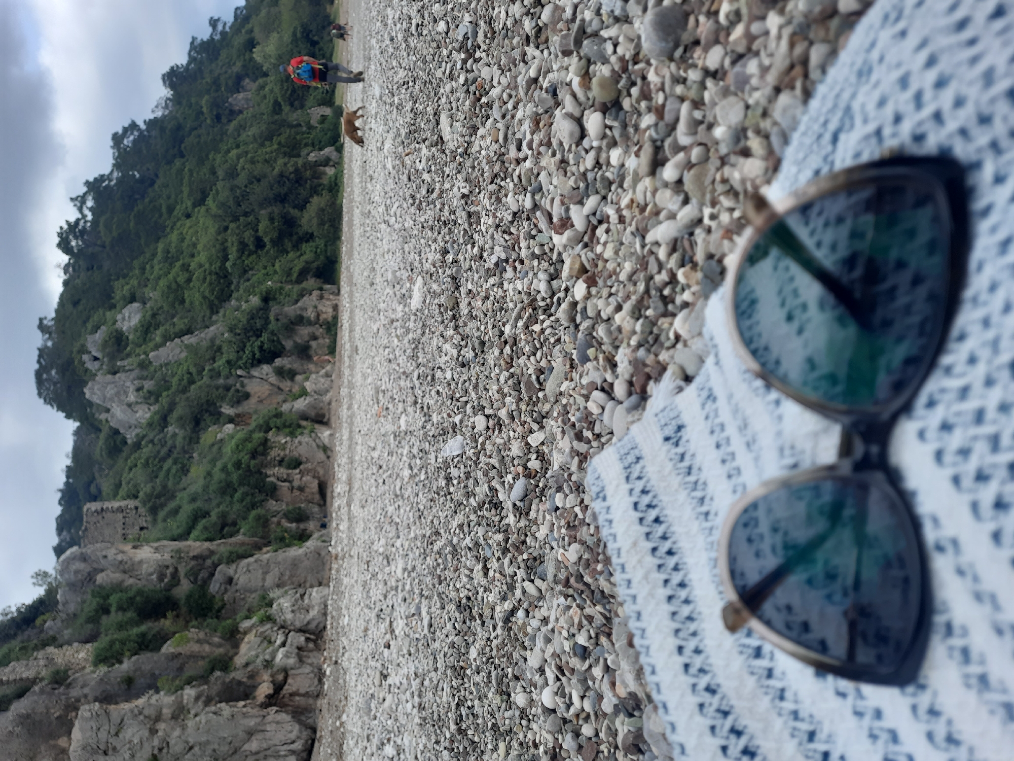 The image captures a scenic pebble beach in Kumluca, Turkey, under an overcast sky. In the immediate foreground, a pair of black-framed sunglasses with dark, reflective lenses is resting on a white textile patterned with blue geometric designs, both slightly out of focus.
The expansive beach is covered with countless light-colored pebbles, stretching into the distance. In the mid-ground, a person is walking on the beach, wearing a red or orange top and a blue backpack. Nearby, a light brown dog is also present. Another indistinct figure, possibly a dog or person, is visible further in the background.
To the left, a steep, rocky hillside covered in dense green trees and vegetation dominates the background. Partially visible on the hillside are ancient stone structures, suggesting ruins or historical fortifications. The sky above is cloudy, indicating an overcast day. No visible text is present.