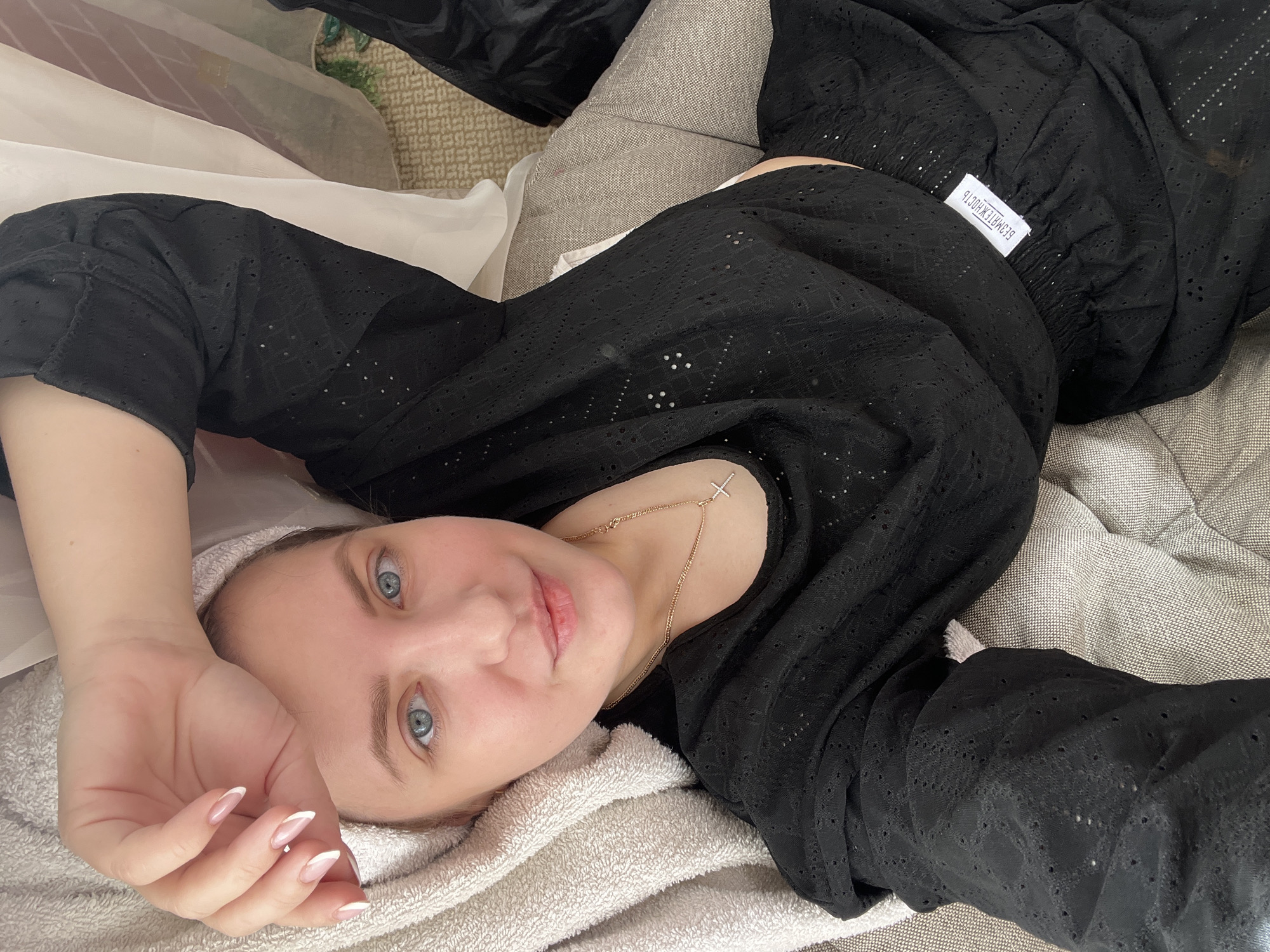 A young woman is lying down, likely indoors. She has fair skin, blue eyes, and light brown hair. She is wearing a black, eyelet-style top, and a gold necklace with a cross pendant is visible. Her nails are painted with a French manicure style. She is resting on what appears to be a white towel or soft cloth, with a pillow or cushion beneath her head. Behind her, there is a beige couch and what looks like dark clothing or material. The woman appears to be looking directly at the camera. The location is Марковский, Russia.