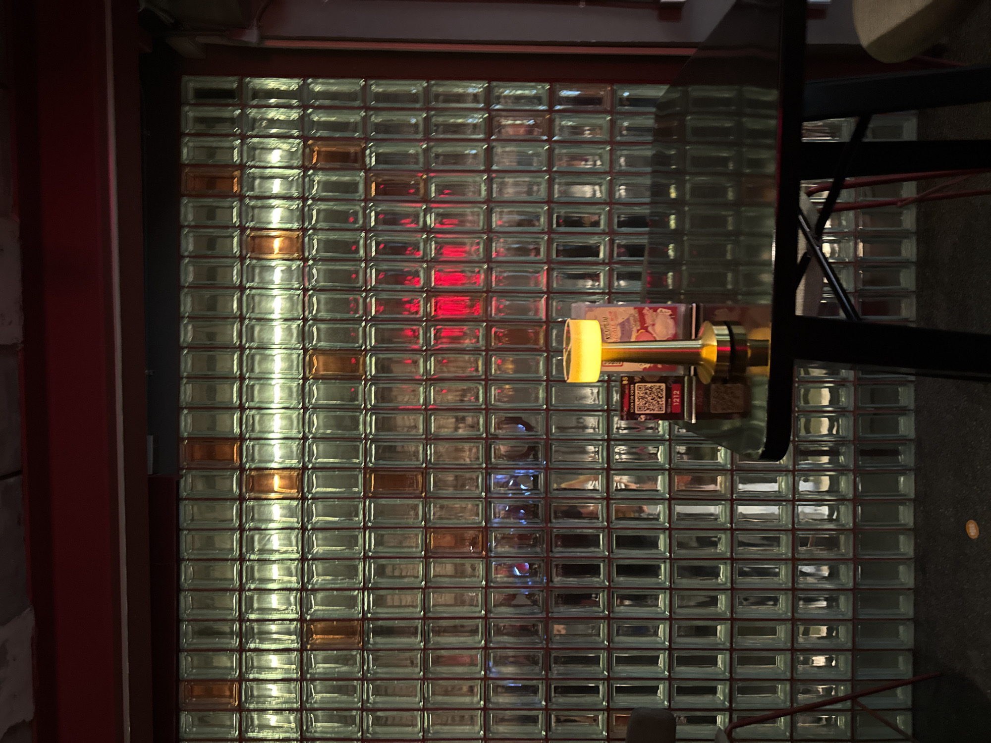 The image depicts the interior of a bar or restaurant in Bangkok, Thailand. The dominant feature is a wall constructed of clear glass blocks, with some blocks exhibiting an amber hue. Red lighting is visible through the glass blocks in the upper portion of the wall, creating a diffused glow. In the foreground, a black, reflective table is situated, with a golden desk lamp and a QR code menu displayed on it. A black chair is partially visible on the left side of the frame, and another red chair is seen in the background. The overall ambiance is dimly lit and intimate, characteristic of a nightlife establishment.