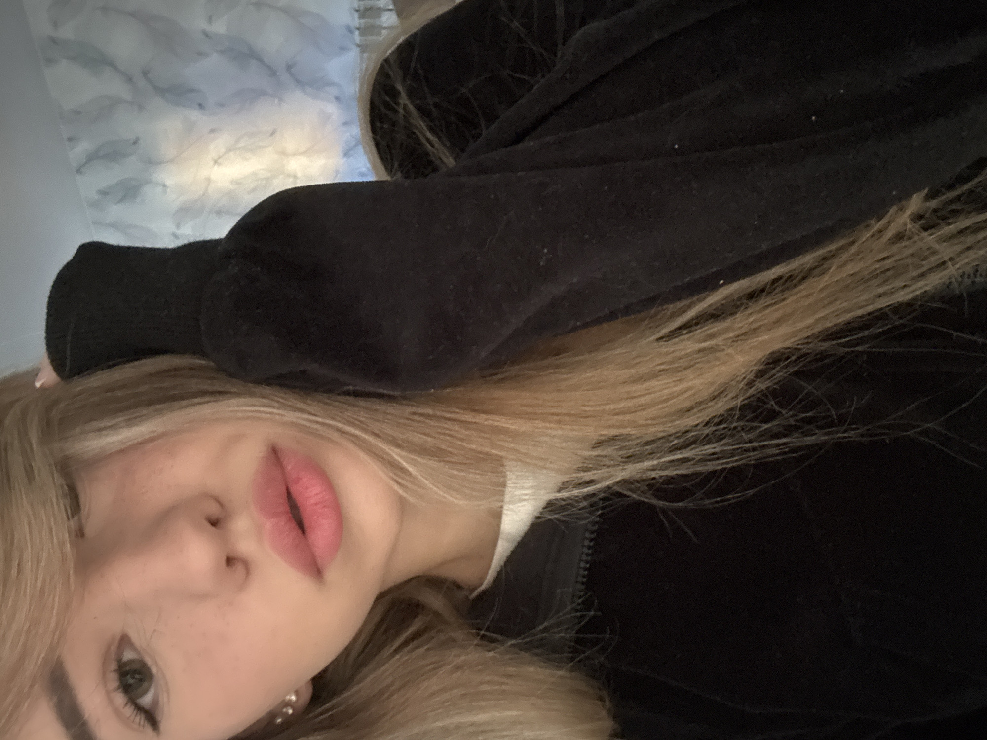 A close-up portrait image, rotated ninety degrees counter-clockwise, features a young person with long, flowing blonde hair and a fair complexion. The person's face occupies the lower left portion of the frame, tilted with their head resting on an arm. Their left eye, enhanced with dark eyeliner, looks towards the viewer. Their lips are slightly parted and adorned with light pink lipstick. Faint freckles are visible across their nose and upper cheeks.
The person is wearing a dark, likely black, hooded sweatshirt or jacket. A segment of its zipper and a patch of white fabric underneath are visible near the neck. A small, round, light-colored earring, possibly a pearl stud, is visible on their left ear. Their right arm, covered by the dark garment, is extended upwards and bent at the elbow, with the hand partially obscured by hair. The fabric of the dark garment shows some small, light specks, possibly dust or lint.
The background reveals an indoor setting with a light-colored wall featuring a subtle, repeating pattern of overlapping, stylized shapes, resembling feathers or leaves. A soft, warm light illuminates the upper right portion of the background. The image captures a posed moment. The provided location context is Noviy, Russia, but no specific visual cues within the image confirm this. There is no visible text.