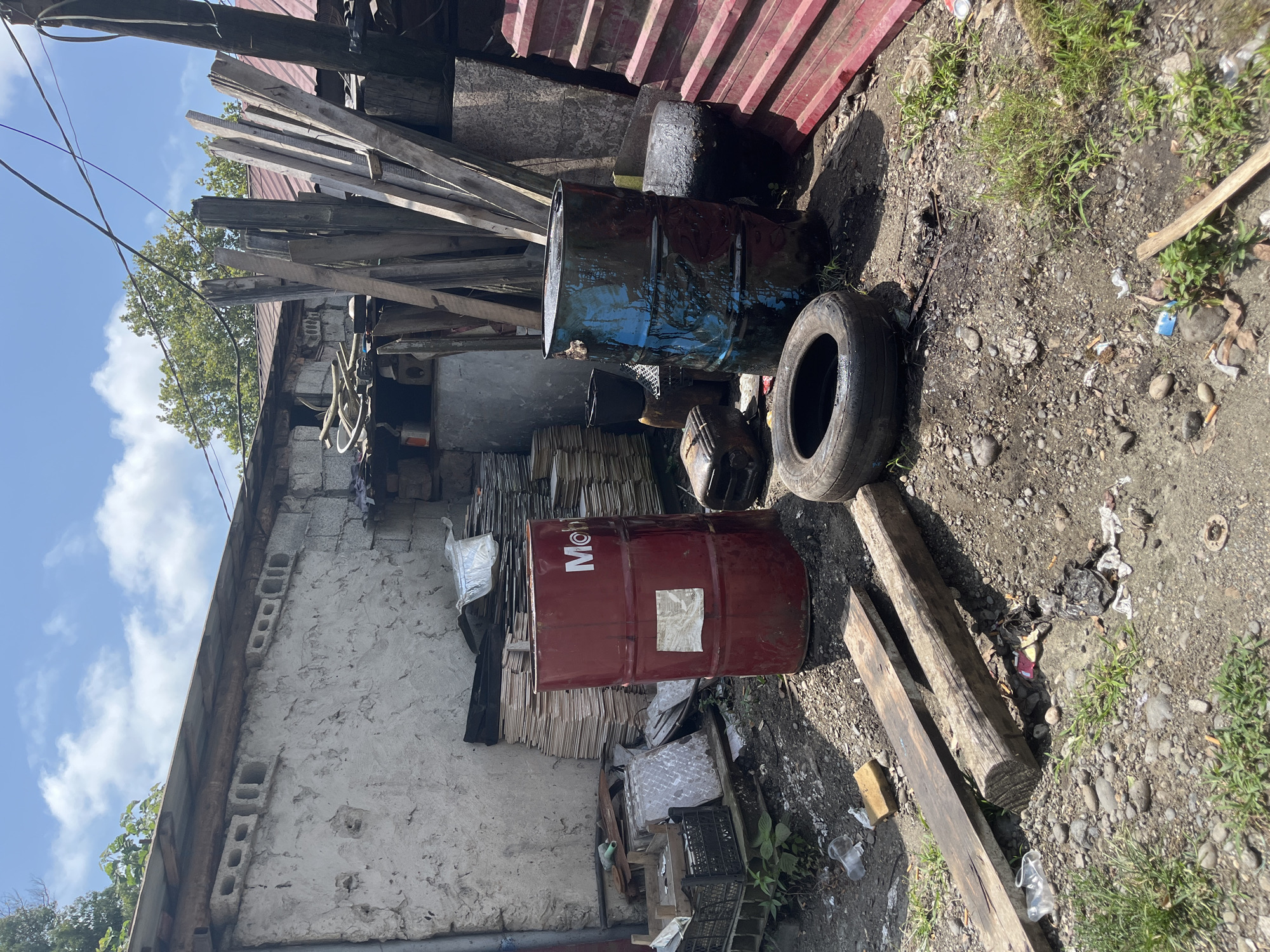 In Ureki, Georgia, an outdoor scene depicts a cluttered area with various discarded items. A red barrel, branded "Mobil," stands prominently in the center, partially obscured by stacks of what appear to be flat building materials. To its right, a dark blue barrel, also cylindrical, is visible, covered in a dark, viscous substance. A deflated tire rests on the ground in front of this barrel.
Behind the barrels, a weathered wall made of concrete blocks is visible, with several rectangular openings or vents. The wall is covered in a rough, off-white stucco or plaster. To the right of the barrels, a stack of weathered wooden planks leans against a structure. In the background, a red corrugated metal fence or paneling is present.
The ground is covered with a mixture of dirt, gravel, and small stones. Scattered debris, including plastic cups, wrappers, and what appear to be cigarette butts, litters the area. Some patches of green grass are growing through the gravel. Overhead, electrical wires are strung across the sky, and a blue sky with scattered white clouds is visible. The scene suggests an area used for storage or possibly a junkyard. No people are present in the image.