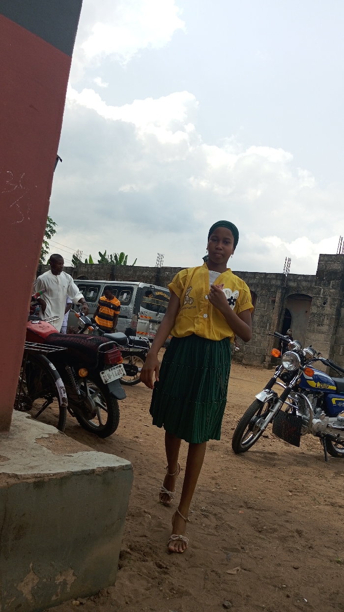 A young woman in a yellow shirt and green skirt walks toward the camera wearing sandals. A red wall is partially visible to the left. Behind her are motorbikes and a van parked in a dirt lot. Two men are visible near the van, and a brick wall stands in the background. The sky is cloudy.
