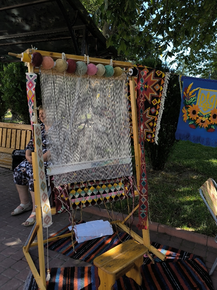 The image depicts an outdoor scene in Illintsi, Ukraine. A large, light-wood upright weaving loom dominates the center, displaying numerous taut white warp threads. A partially woven section near the bottom features horizontal geometric patterns in black, pink, yellow, green, and light blue. Nine yarn balls in varied colors (yellow, maroon, light pink, light green, dark green, dark pink, light blue, purple) are suspended from the loom's top bar. A narrow, vertically oriented woven band with geometric designs hangs from the loom's left support.
To the left of the loom, a light-skinned adult female, dressed in a dark patterned top and light sandals, is seated on a slatted wooden bench, facing forward with a slight smile. She is not actively engaged with the loom. To the right of the loom, a finished black-and-red patterned rug hangs vertically. Further to the right, a blue tapestry depicting the Ukrainian Tryzub emblem surrounded by sunflowers is displayed. A small wooden stool is positioned on a striped woven rug in front of the loom. A stack of white papers or fabric rests on a lower crossbar of the loom. The ground is paved with red-brown interlocking bricks. Green trees and foliage are visible in the background under a blue sky, with a dark metal pergola structure partially visible in the upper left. The scene is illuminated by bright natural daylight.