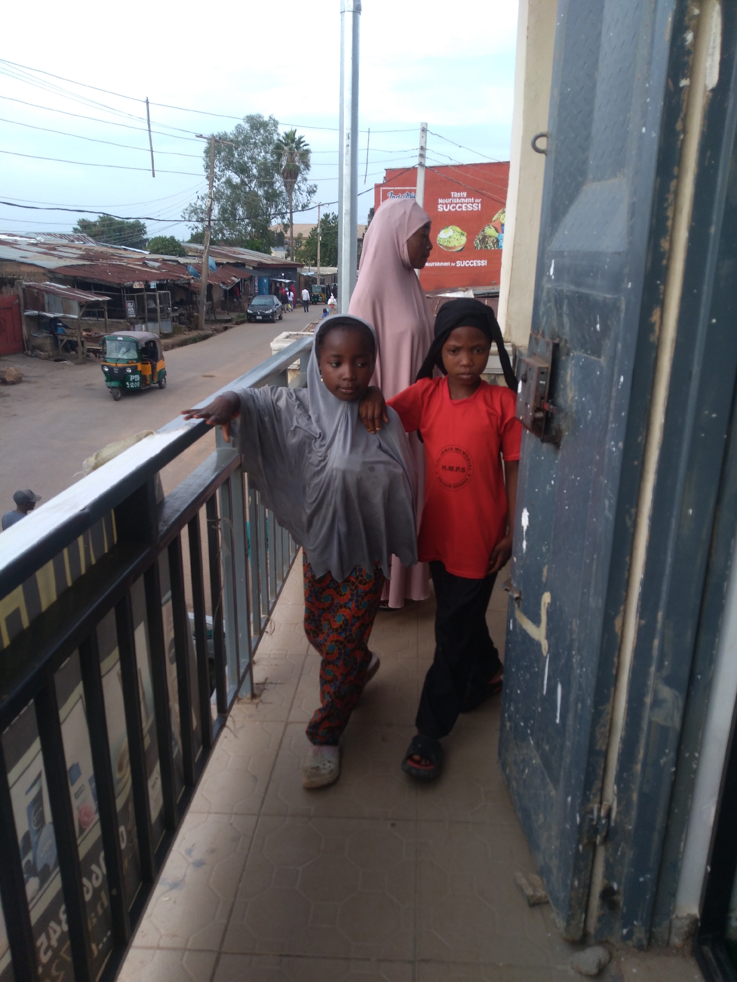 This image captures two young girls standing on a balcony or raised walkway overlooking a street scene in Jos, Nigeria. The girl on the left wears a grey hijab and a patterned skirt, while the girl on the right sports a black headscarf and a red t-shirt with "H.M.P.S." visible in a circular emblem. They appear to be posing for the photo, with the girl on the left resting her hand on the railing.
Behind them, a woman in a light pink hijab is partially visible, suggesting they are together. The street below is lined with small shops or market stalls with corrugated iron roofs, indicating a bustling commercial area. A three-wheeled auto-rickshaw, commonly known as a "keke" in Nigeria, with license plate "5208" is visible on the street. Further down the road, a few cars and people are present, hinting at an active daytime scene.
The sky is overcast, suggesting a cloudy day. The overall impression is a snapshot of everyday life in a Nigerian urban setting, focusing on the young subjects in the foreground against a backdrop of local commerce and daily activities. The visible text includes brand names and what appears to be school or organization affiliation, adding a layer of cultural context.