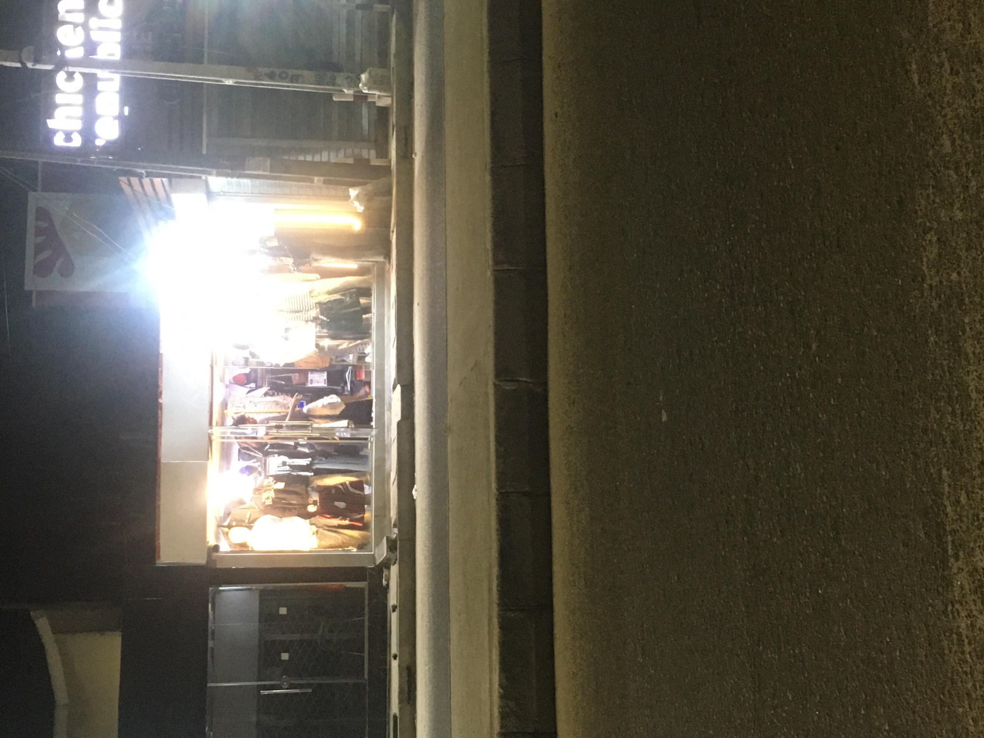 The image depicts a storefront at night. The sign above reads "Chicken Republic." Inside the store, there is a display case filled with items. People are visible inside the store. The location is likely in Ado, Nigeria.