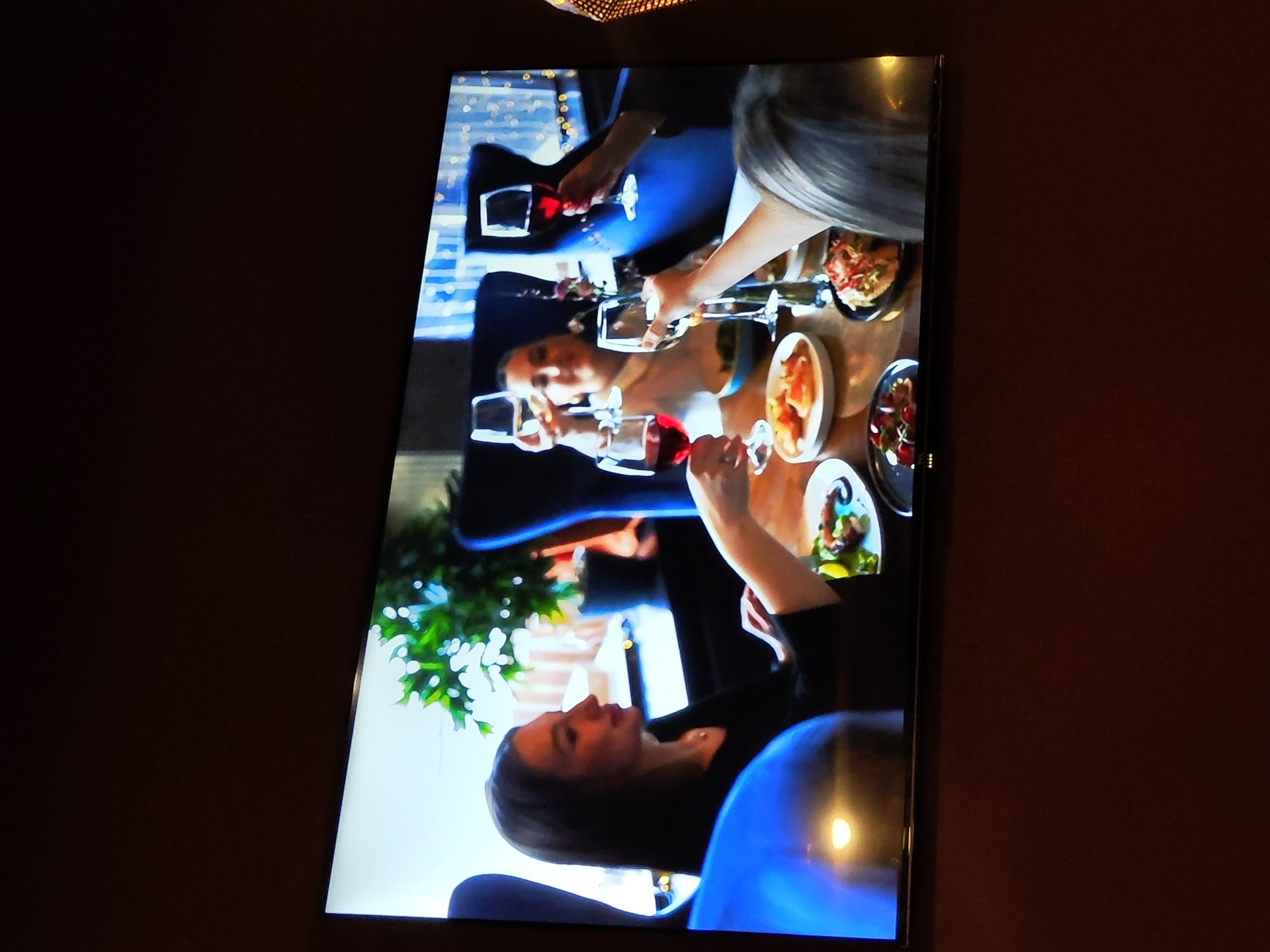 The image shows a television screen displaying a scene of people dining. In the foreground, a woman in a black top is seated at a table, holding a glass of red wine. In the background, three other individuals are visible, also holding wine glasses. The table is set with several plates of food, including appetizers. The environment appears to be a restaurant setting, with plush seating and decorative lights. The television is mounted on a dark brown wall. The image does not contain any information that would indicate a specific location in Zhukovsky, Russia.
