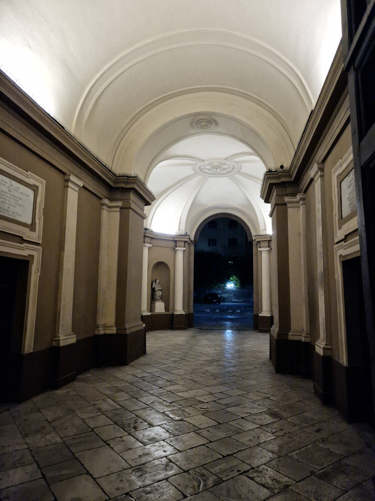 This image shows an illuminated arched passageway with a stone-tiled floor. The passageway features classical architectural elements such as columns and a decorative ceiling with a central medallion. On the left wall, there is a framed plaque with text. Further down the passageway, a small statue is visible in an alcove. The far end of the passageway opens onto a dark street with a building visible in the distance, illuminated by some lights. The floor appears wet, suggesting recent rain. The overall scene depicts a historic architectural structure in an urban environment at night.
This is a view of the passage leading to the courtyard of the former Seminario Maggiore Militare (Major Military Seminary) in Taranto, Italy. The building is located at Via Duomo, 200.