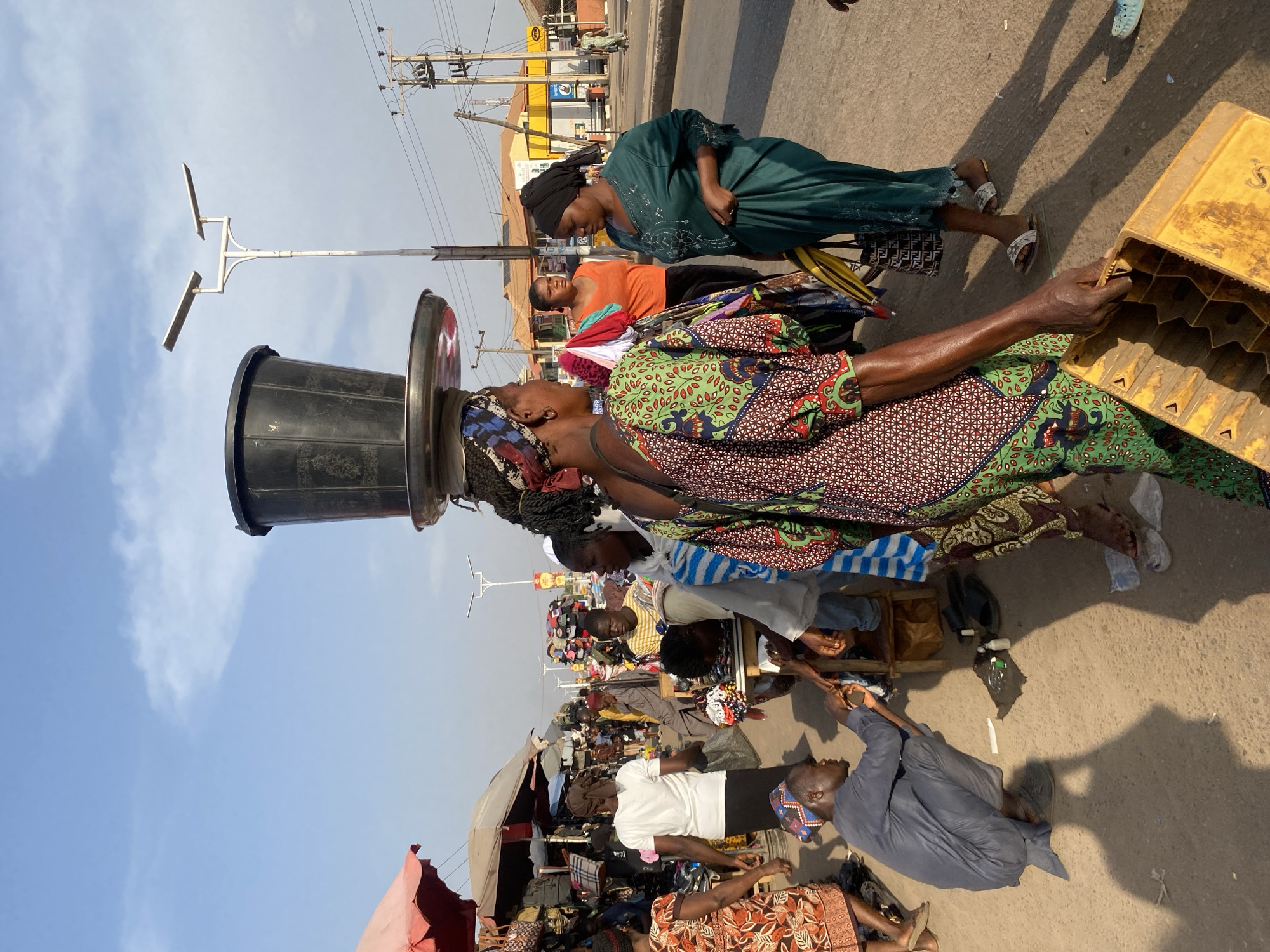 The image depicts an active outdoor market scene on a sunny day in Sabon Tasha, Nigeria. The foreground is dominated by a woman walking towards the viewer's right, balancing a black bucket on a silver tray on her head. She wears a colorful patterned dress and carries a yellow plastic crate in her right hand. Her hair is braided and partially covered by a multi-patterned headwrap. To her immediate right, a woman in a dark green embellished dress faces left, holding a yellow item and wearing white and black patterned slides. Behind the central figure, another woman in an orange shirt stands facing right.
In the mid-ground, a man in a blue kaftan and patterned cap squats on the unpaved ground, appearing to engage with small objects or tools placed before him, including bottles and a dark cloth. Nearby, market stalls with red and white umbrellas display various goods such as bags, shoes, and hats. Several other individuals are present, including a woman in a white t-shirt walking away from the viewer, carrying a black bag, and a man in a striped shirt seated behind a display of hats.
The background reveals a street with utility poles carrying electrical wires and a white street light pole with two horizontal fixtures. A yellow building with a red roof is visible further down the street. The ground is light-colored and appears dusty or unpaved, with long shadows cast, suggesting strong daylight. The sky is clear with scattered white clouds. Multiple people are scattered throughout the scene, indicating a bustling commercial environment.