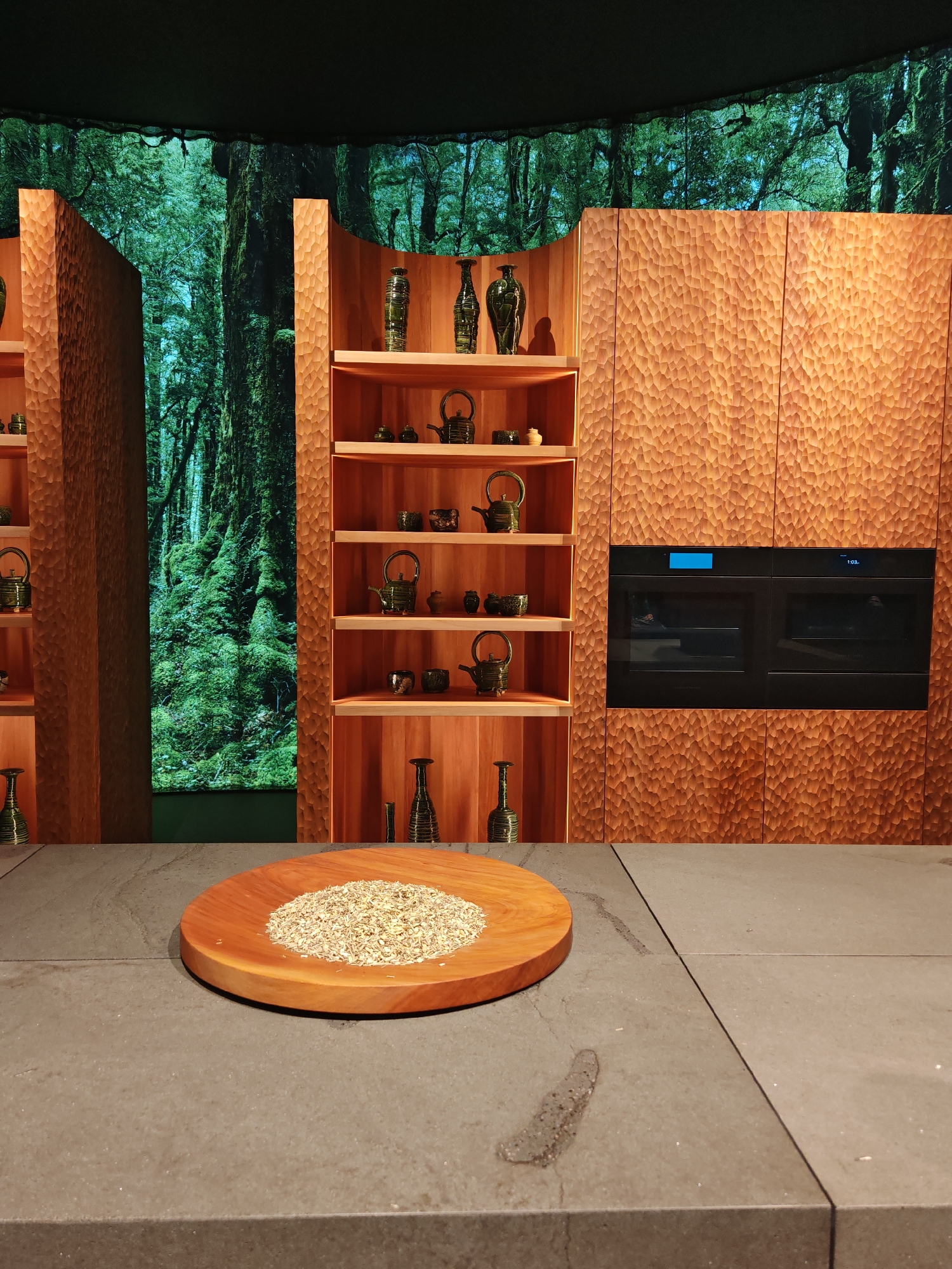 The photograph shows a kitchen in Unknown, Unknown. The counter, made of matte gray material, displays a round wooden cutting board containing a pile of light brown shredded material. To the rear is a backdrop with a green forest scene. The right side of the counter is an adjacent counter of the same material. Behind the counter are a built-in wall oven and shelving, both constructed of a brown wood with a carved texture. The shelving holds various decorative pottery teapots and vases.