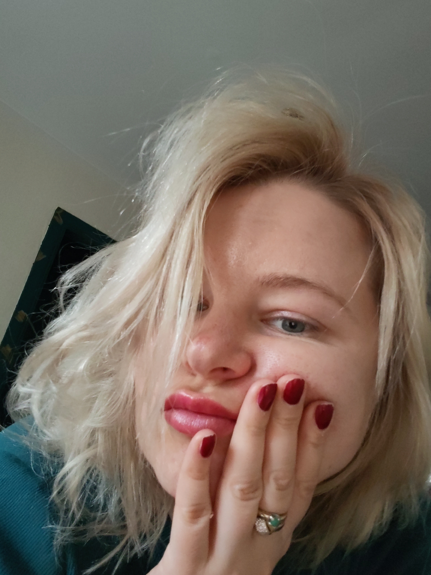 A close-up, eye-level shot shows a fair-skinned woman with blonde, slightly messy hair. Her hair is styled with some strands falling across her face. Her eyes are blue. She is pursing her lips in a pout. She has red nail polish on her fingernails. She wears a ring on her ring finger. Her left hand is against her cheek. She is wearing a teal shirt. The background is a plain wall, with a dark, framed object visible in the upper left corner. There is no information to confirm that the location is in Izhevsk, Russia.
