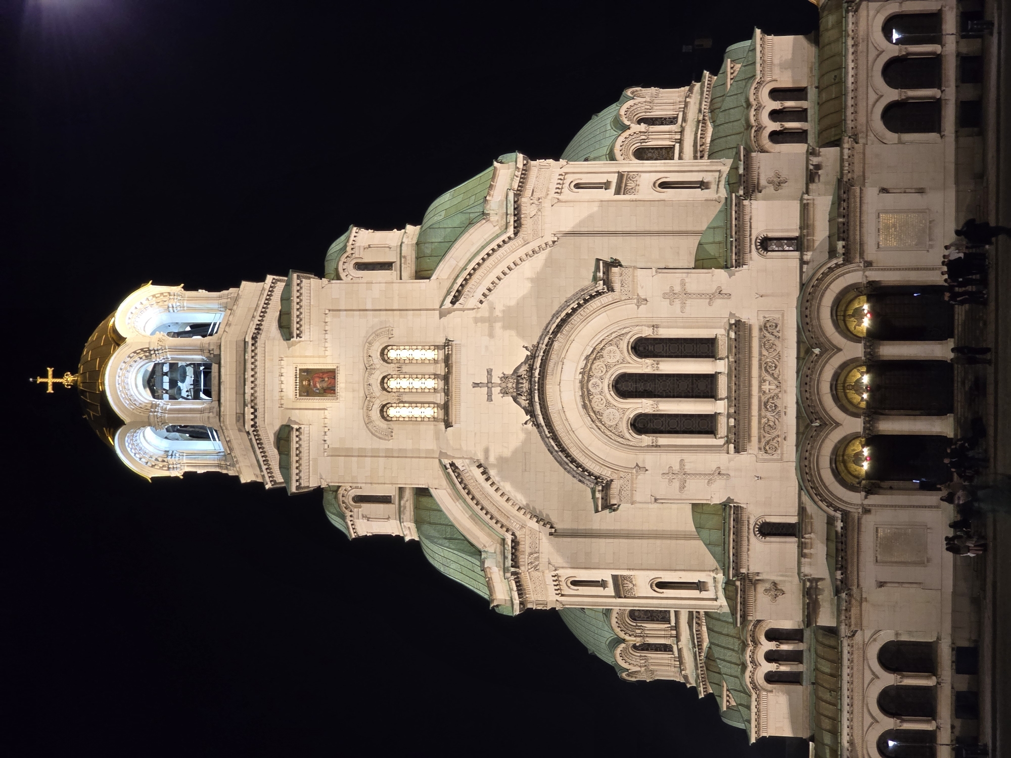 This image captures a majestic, ornately designed cathedral illuminated at night against a stark, dark sky. The architectural marvel, consistent with the Alexander Nevsky Cathedral in Sofia, Bulgaria, stands prominently, showcasing its impressive scale and intricate details.
The structure features multiple domes, with a striking golden dome crowned by a golden Orthodox cross positioned towards the upper left. Several other domes are visible, characterized by their distinctive greenish-blue patinated surfaces. The building's facade is a rich tapestry of intricate stone carvings, including decorative arches, columns, pilasters, and friezes. Numerous arched windows, some glowing with interior light, are interspersed across the stonework. A belfry, located beneath the golden dome, reveals large bells within its arches.
Religious iconography is present in the form of small, colorful mosaic or fresco-like images depicting figures on the upper sections of the walls. Towards the lower right of the image, a group of several people can be seen standing near the base of the building, providing a sense of scale and indicating a public setting.
The time of day is clearly night, with the building brightly lit by artificial sources that accentuate its grandeur and textures. The sky is black and clear, with only a very faint light visible in the top-left corner, possibly a distant light or celestial body. A light-colored rectangular plaque with unreadable text, likely in Cyrillic, is visible on the right side of the building's facade. The scene evokes a sense of awe and the historical and cultural significance of the monument.
