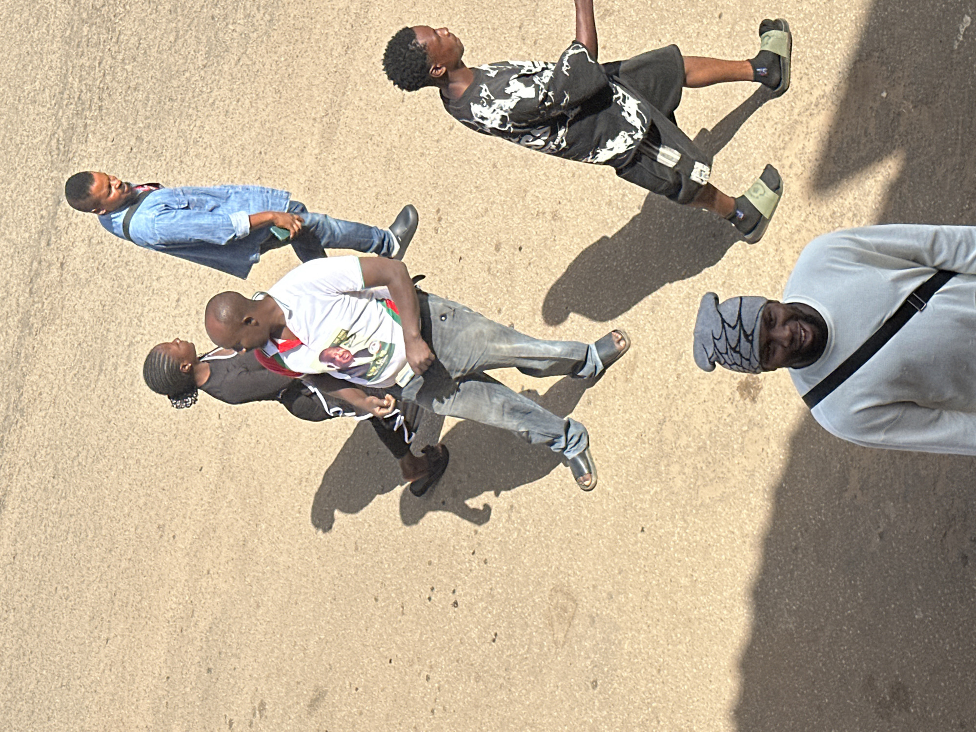 An aerial shot captures five individuals walking across a sun-drenched, sandy street in Bukuru, Nigeria. In the foreground, a Black man wearing a gray beanie and a light-colored long-sleeved shirt with a black strap across his chest looks at the camera with a slight smile. Behind him and to the right, a young Black man in a black and white patterned t-shirt and black shorts walks away from the camera, wearing dark socks and light-colored slides. Further back and to the left, a Black man in a white t-shirt with a political image on it, dark gray jeans, and black sandals walks away from the camera. To his left, a Black woman with braided hair, wearing a black t-shirt and black pants, walks away from the camera. In the background, a Black man in a light blue denim jacket, blue jeans, and black slides walks away from the camera, holding a green object in his right hand. The ground is a textured, sandy beige, with scattered dark specks. Shadows are cast long and dark across the street, indicating the bright sunlight.