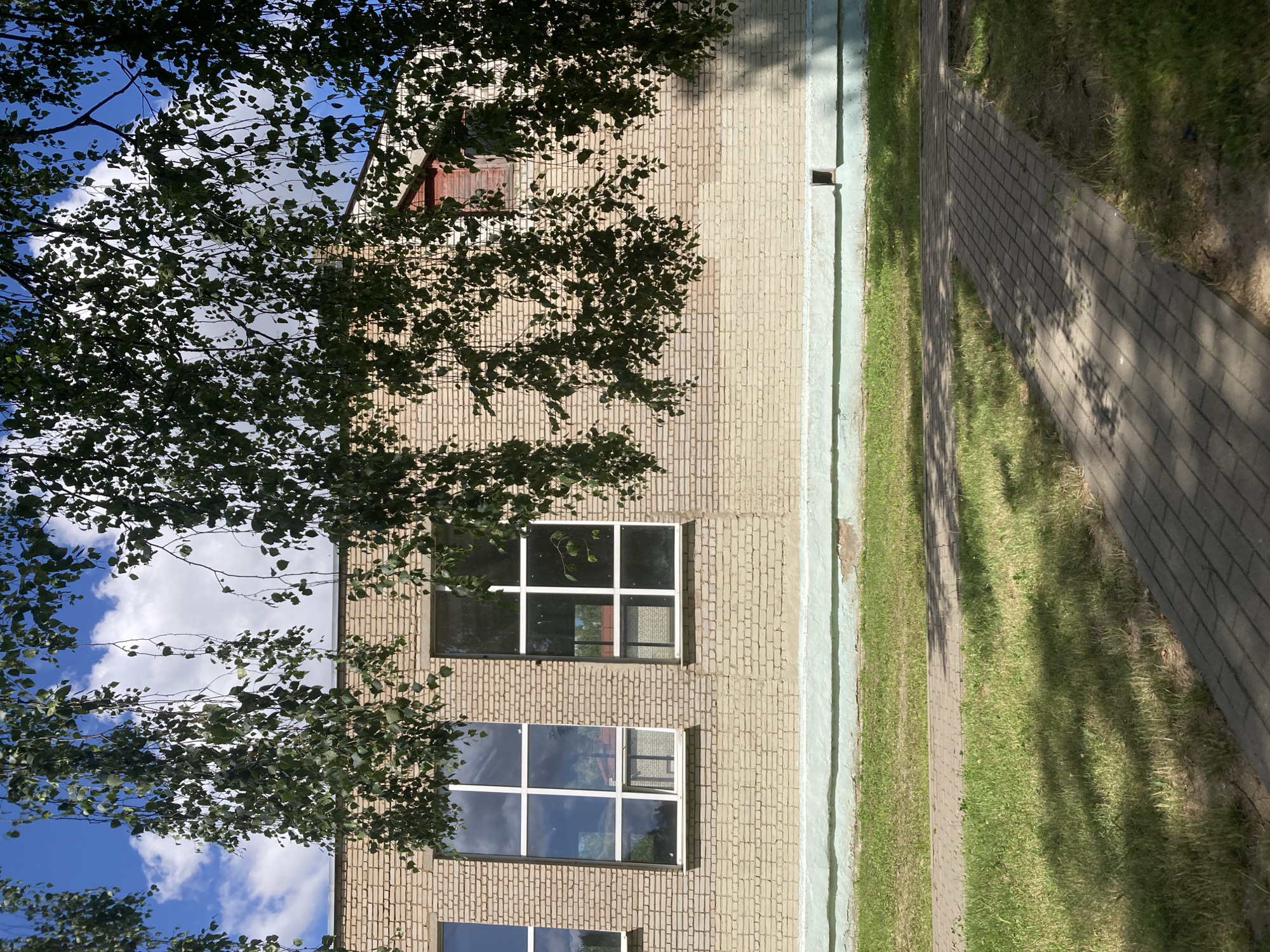 The image shows a brick building with large windows. The building is likely a school or public institution, with a green painted strip at the base and a sidewalk leading away from it. The sky is blue with white clouds, and trees with green leaves hang over the building. The scene is set in Zhodzina, Belarus. There are no people or animals visible in the image. The focus is on the architecture of the building and its surroundings.