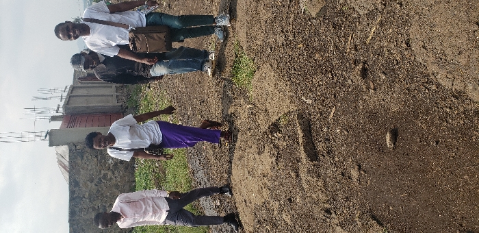 The image shows four individuals walking on an unpaved dirt path in Buzove, Democratic Republic of the Congo, under an overcast sky. From left to right: a male in a light-colored collared shirt, dark trousers, and dark shoes; a female in a white t-shirt, purple trousers, and sandals, carrying a dark shoulder bag; a male in a dark shirt, blue jeans, light athletic shoes, and a dark baseball cap; and another male in a white t-shirt, blue jeans, and light athletic shoes, carrying a brown shoulder bag and holding papers in his left hand. The female and the rightmost male are smiling. The ground is composed of loose brown soil, uneven with slight mounds and depressions, and contains sparse green vegetation. In the background, a low stone wall, a red-brown metal gate, and unfinished concrete structures with exposed rebar are visible. Distant terrain or structures are faintly discernible on the horizon to the far right.