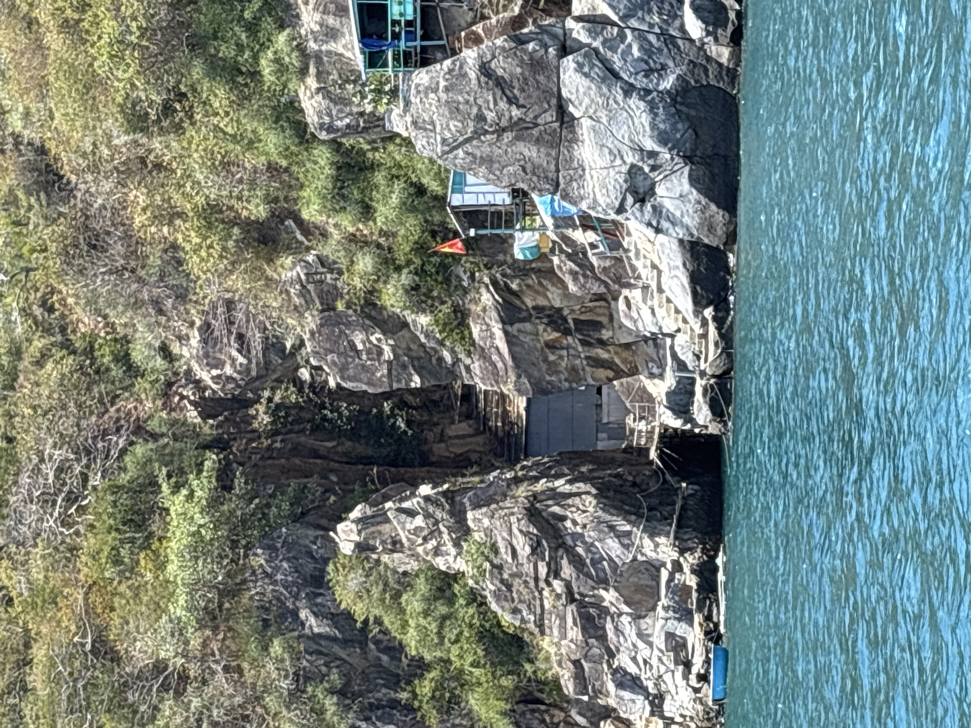 The image depicts a steep, rocky cliff face covered with vegetation in Bac Nha Trang Ward, Vietnam. At various points on the cliff, small structures are built into the rock. One structure appears to be constructed from metal framing and has a blue awning. Another has a small orange flag. The cliff meets the edge of a body of water to the right of the frame. The water is turquoise in color and appears calm.