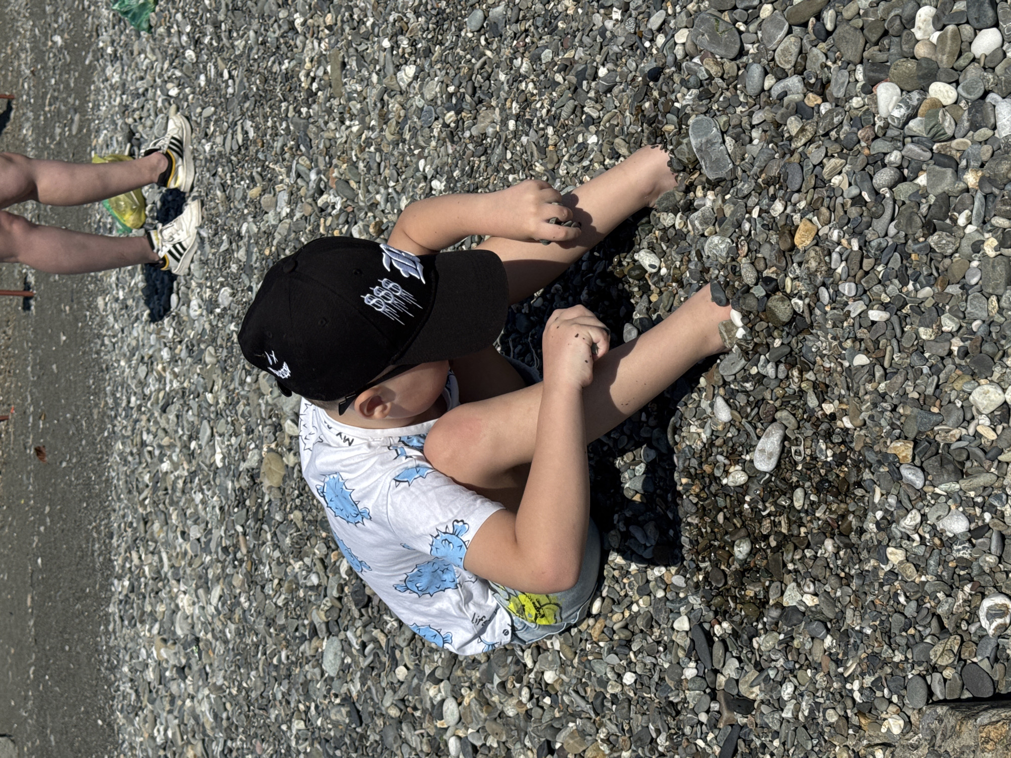 A young child in a black baseball cap with a white graphic, a white t-shirt with blue fish designs, and light-colored shorts sits on a beach covered in small, gray and white pebbles. The child is using both hands, appearing to manipulate the pebbles. To the left, a pair of legs wearing Adidas sneakers is visible. The location appears to be a pebbled beach near the sea, possibly in Sochi, Russia.