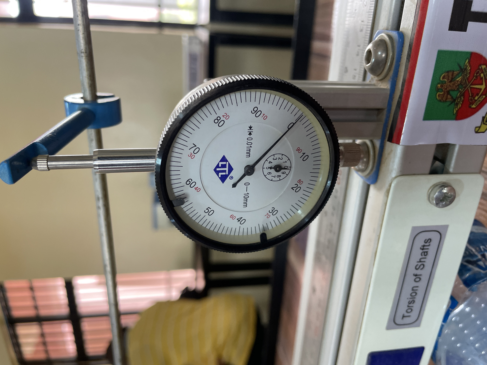 The image shows a close-up of a dial indicator gauge, which is a precision measuring instrument. The needle on the main dial points between "80" and "90". The smaller dial indicates a value around "5". The dial indicator is attached to a metal frame, and a thin metal rod extends from the gauge, presumably to make contact with the object being measured. A label on the frame reads "Torsion of Shafts". A flag with a heraldic emblem is visible on the frame. The dial indicator is placed somewhere in Afaka, Nigeria.