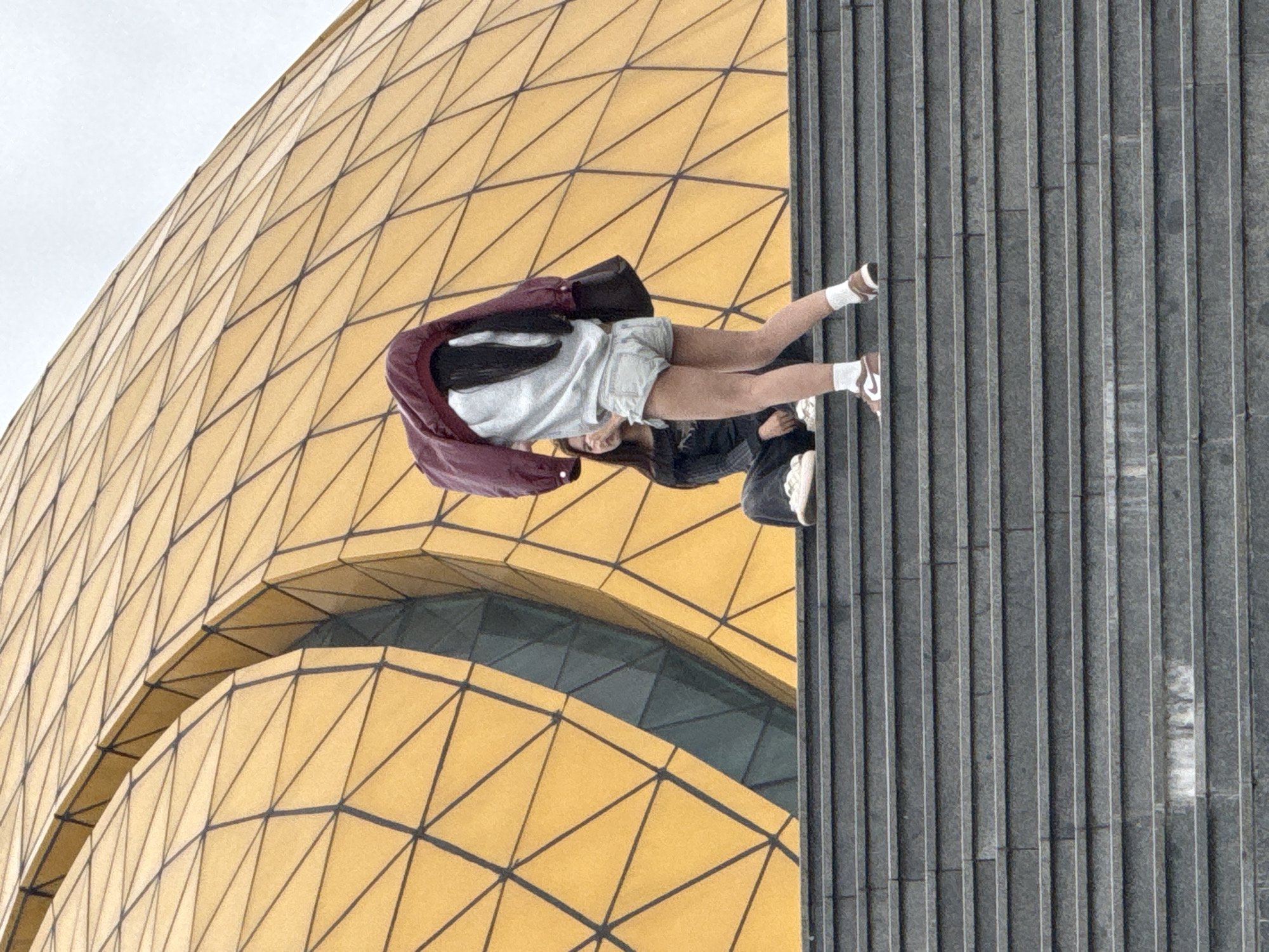 The image depicts two individuals on a wide outdoor staircase leading up to a striking golden, geodesic dome-like structure. The dome's surface is characterized by a repeating pattern of triangular panels, creating a honeycomb or diamond-like appearance. The staircase itself is constructed of dark gray stone or concrete, with distinct horizontal steps.
In the foreground, a person is standing on the stairs, facing away from the camera and towards the dome. This individual is wearing a light gray sweatshirt, denim shorts, white socks, and brown and white sneakers. A dark red jacket is draped over their shoulders, obscuring their face and hair. A dark bag is slung over their left shoulder.
Further down the stairs, to the left and slightly behind the standing individual, another person is seated. This person is wearing a dark long-sleeved shirt and dark pants, with white sneakers. They are looking towards the person in front of them.
The background is dominated by the intricate golden facade of the dome and a hazy, overcast sky. The overall scene suggests a public space, likely a tourist attraction or landmark in Da Lat, Vietnam, given the architectural style. The lighting is diffused, indicating a cloudy day.