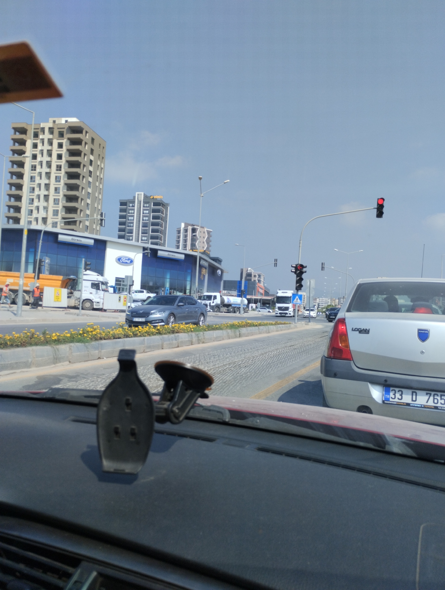 The image captures a street scene in Mezitli, Turkey, likely along a commercial strip. In the foreground, the interior of a car is visible, with a dashboard-mounted phone holder prominently displayed. The perspective is from the driver's seat, looking out onto the road.
Outside the car, a multi-lane road stretches into the distance, lined with yellow flowers along a median barrier. On the left side of the road, a modern Ford dealership with a blue facade and large windows is visible. A white truck with an orange dumpster, and two construction workers in orange vests, are parked in front of the dealership. Several other vehicles, including a gray sedan and a silver Dacia Logan, are on the road.
In the background, several tall buildings, characteristic of urban development, rise against a bright blue sky with scattered clouds. Traffic lights are visible, with one displaying a red light. The overall impression is a sunny day in a developed urban area with ongoing commercial activity and traffic.