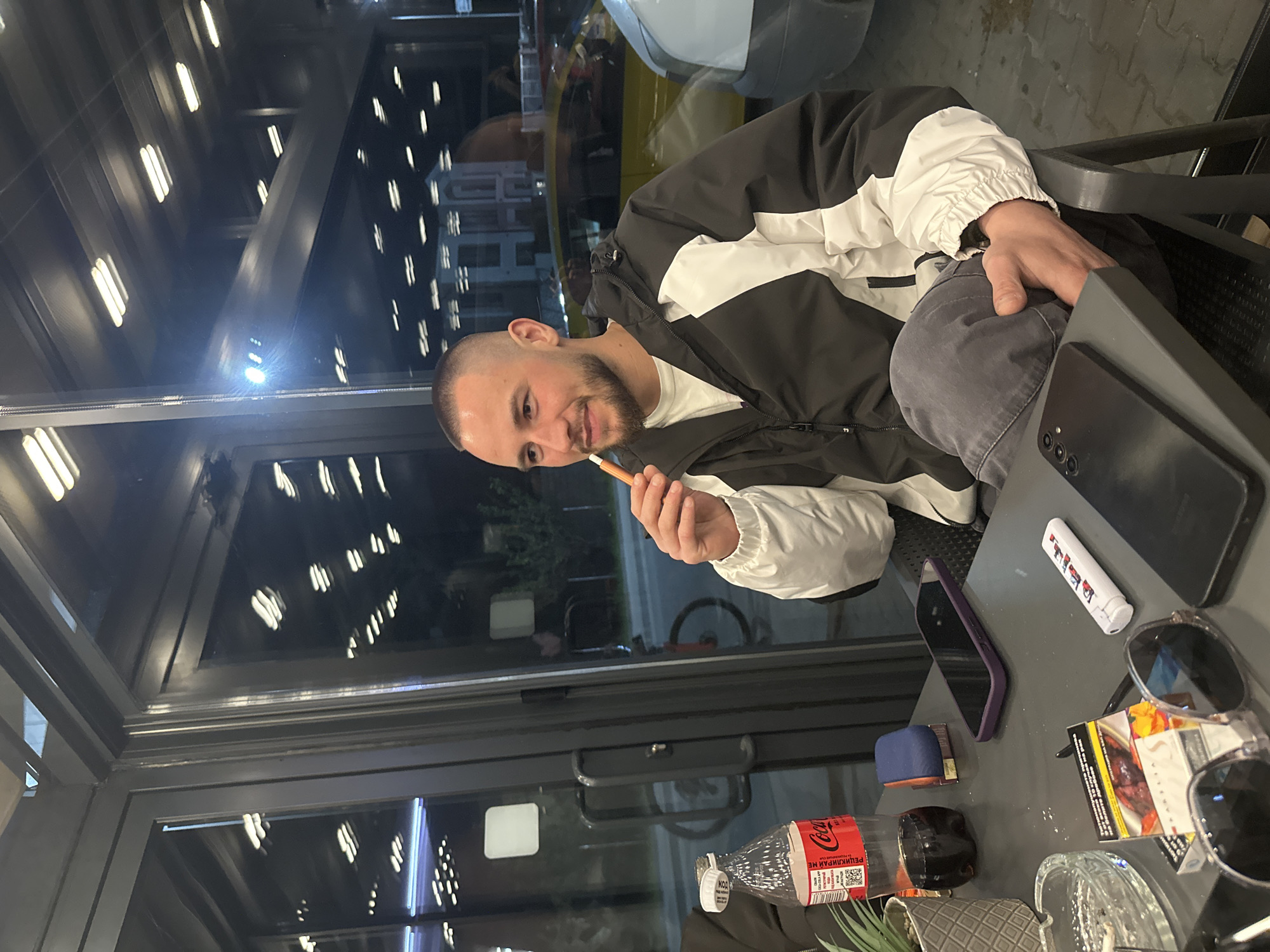The image displays a young man seated at a table within what appears to be a cafe or restaurant during the evening or night. He is the central subject, featuring short hair, a trimmed beard, and a slight smile. He wears a black and white windbreaker jacket over a white t-shirt and grey pants, holding a slender, light brown object, likely a vape or e-cigarette, near his mouth.
The setting includes large glass windows in the background that reflect bright, linear indoor lighting fixtures, indicative of nighttime. Through these reflections, subtle hints of an urban environment, including buildings and a vehicle, are visible, along with a bicycle further in the distance. The man is sitting on a dark chair at a dark, reflective table.
On the table, several items are arranged: a black smartphone, another smartphone in a purple case, a white lighter with red markings, a pack of cigarettes, clear-framed sunglasses, a glass ashtray containing a cigarette butt, and a plastic Coca-Cola bottle. A small blue object and a partial view of a potted green plant are also present.
The Coca-Cola bottle clearly displays "Coca-Cola" and "РЕЦИКЛИРАЙ МЕ" (Recycle Me) in Cyrillic script. The cigarette pack also features Cyrillic text for health warnings. These linguistic cues, along with the urban environment, are consistent with the provided location context of Plovdiv, Bulgaria. The man's relaxed posture suggests a moment of leisure or socializing.