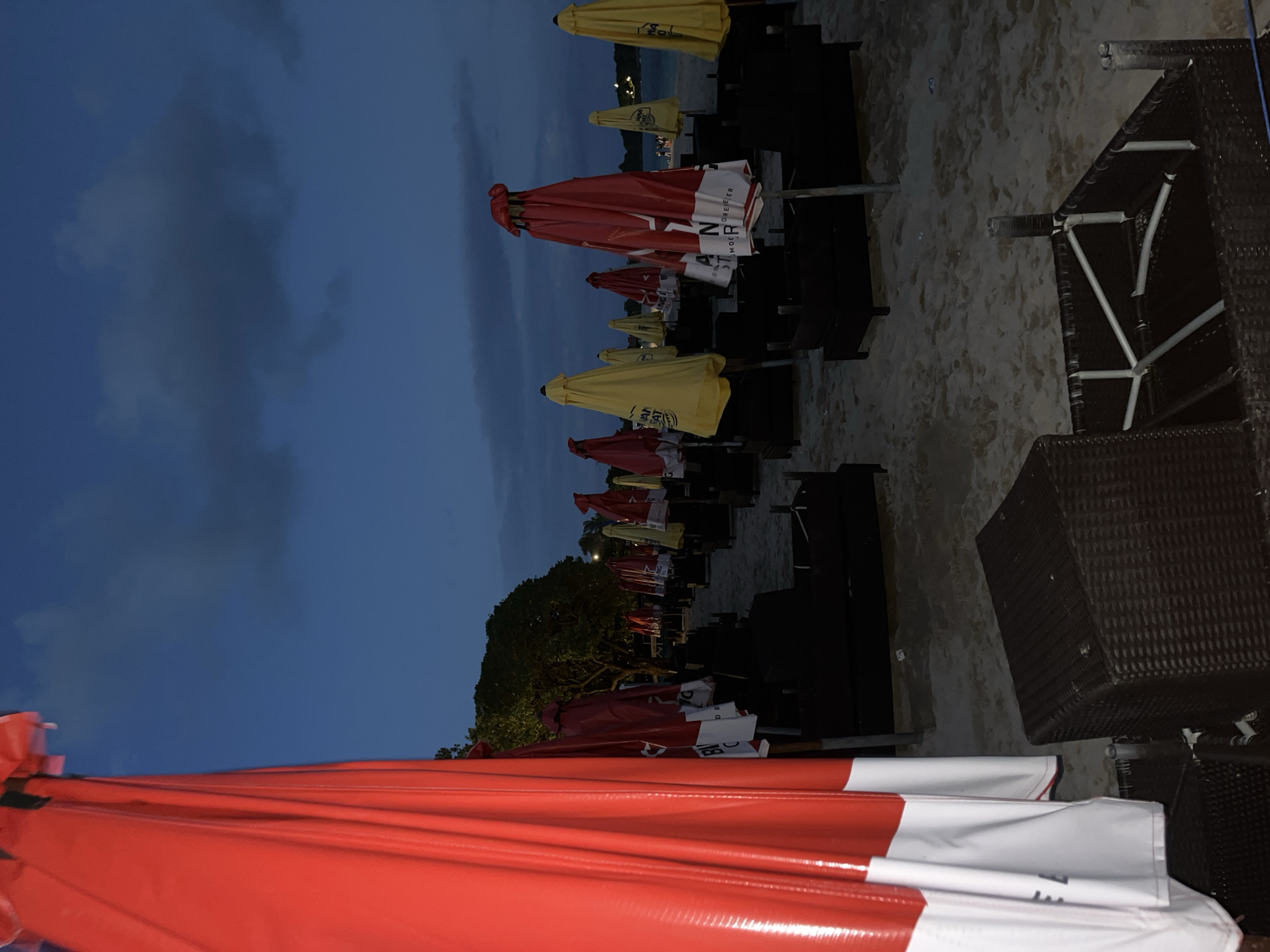 The image shows a row of folded beach umbrellas and lounge chairs on a beach in Kuta Selatan, Indonesia. The umbrellas are primarily red and yellow. The sky is dark blue, indicating either evening or approaching rain. A line of trees is visible in the middle of the image. The ground is sandy.