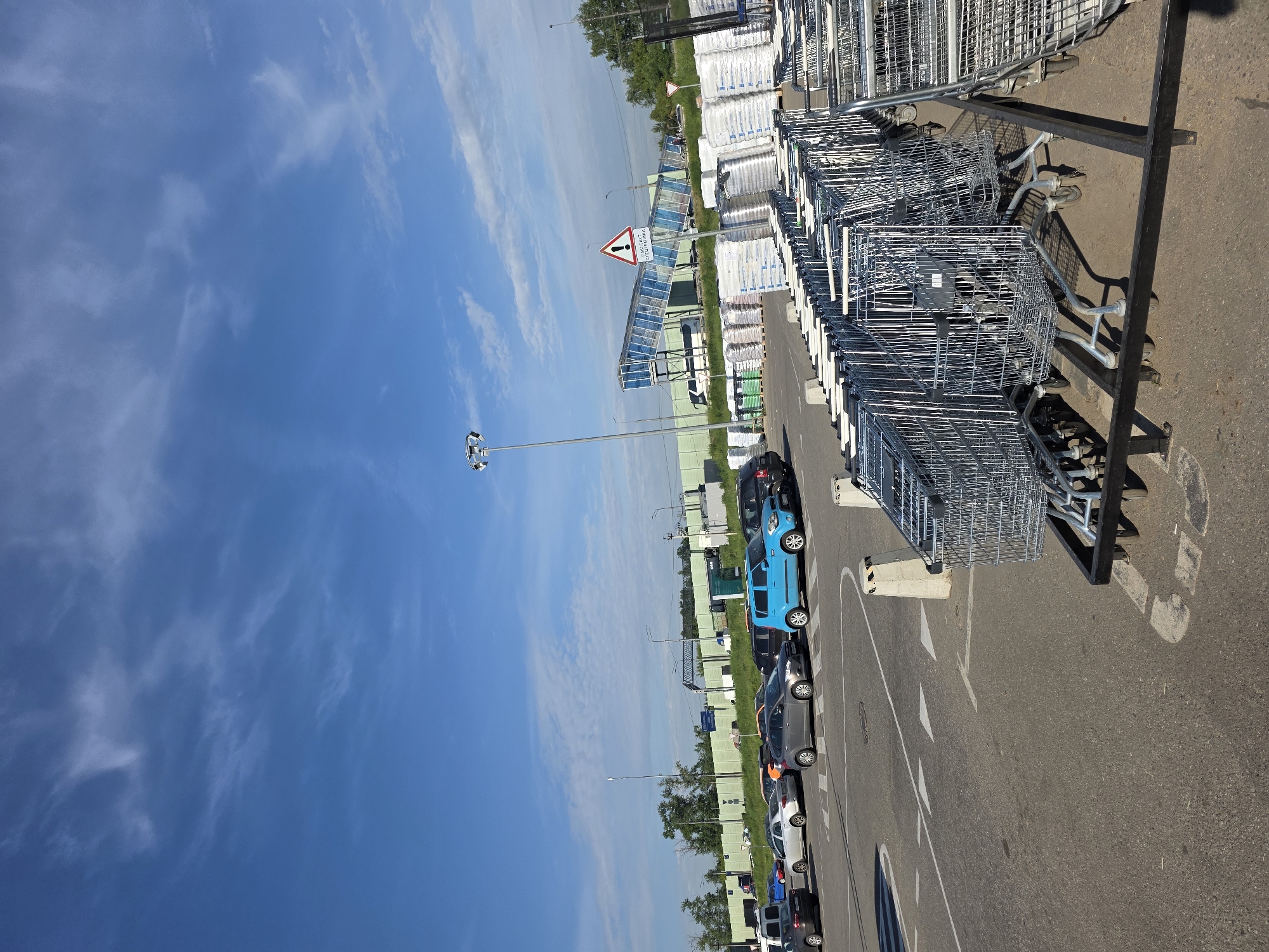 A low-angle, wide shot captures a parking lot filled with shopping carts, cars, and a bright blue sky with scattered clouds. In the foreground, rows of metal shopping carts are neatly arranged on black metal racks. Beyond the carts, a paved parking lot is marked with white lines, and several cars are parked or driving. A bright blue car is prominent among them. In the middle ground, a long, green fence runs horizontally, with a pedestrian overpass with blue tinted glass and metal construction spanning over it. The overpass has a warning sign with an exclamation point and Russian text. To the right of the overpass, stacks of white, bagged materials are piled high. The background shows a line of trees and a bright blue sky with wispy white clouds. The overall scene suggests a commercial area, possibly a large retail store with an outdoor parking lot and delivery area.