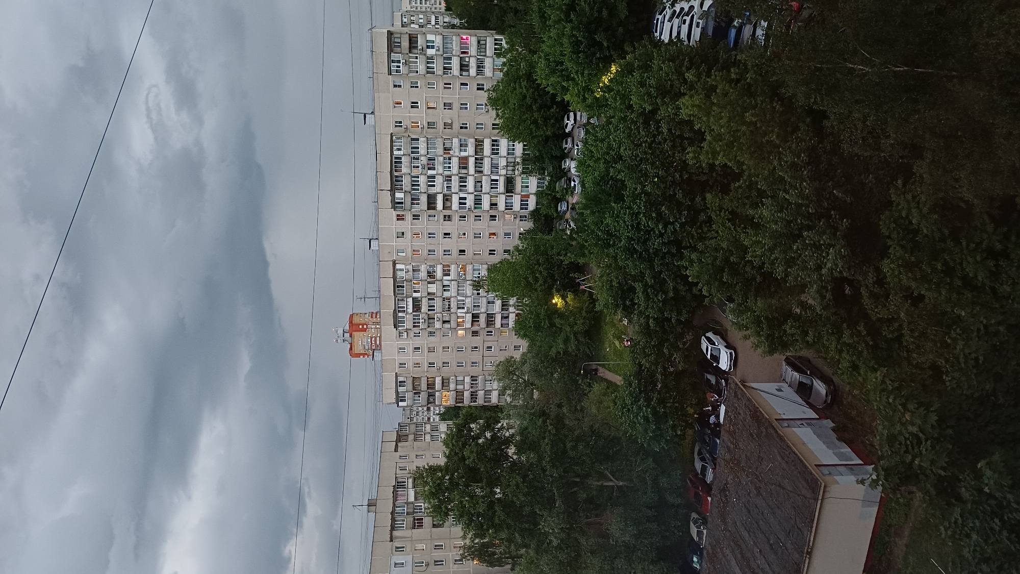 This is an exterior, medium-shot photograph taken in Balashikha, Russia. The image shows an urban landscape with tall apartment buildings, trees, and a cloudy sky. The buildings are typical Soviet-era residential blocks. In between the buildings are green trees. There are parked cars near the buildings. The sky is overcast.