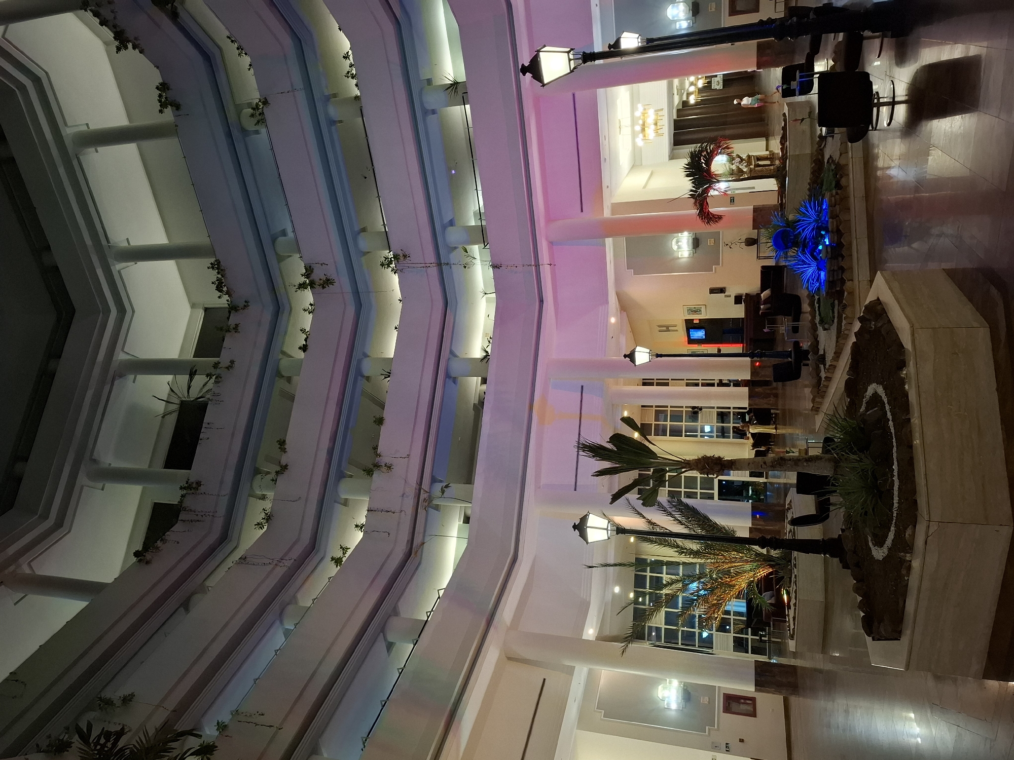 The image shows the interior of a multi-story building, likely a hotel or resort, in Pedro González, Venezuela. The architecture features tiered balconies lined with plants, creating a vertical garden effect. The color palette is predominantly white and beige, illuminated by artificial lighting with pink and blue hues. On the ground floor, there are potted plants, decorative lighting fixtures resembling antique street lamps, and seating areas. The floor is tiled. The scene is devoid of people.