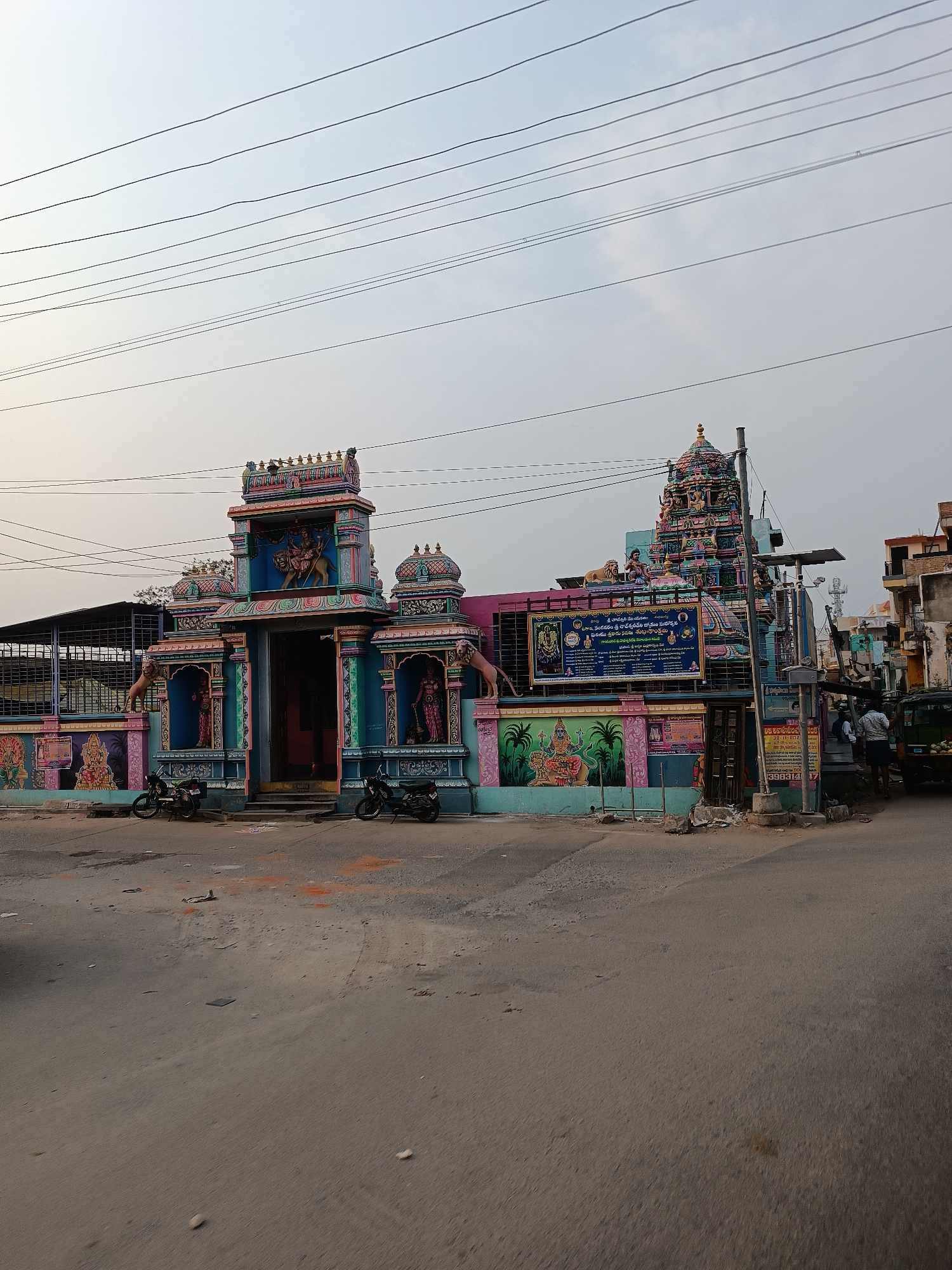 The image depicts a colorful Hindu temple located in what appears to be a city in Dharmavaram, India. The temple is adorned with vibrant paintings and sculptures of deities. Two motorcycles are parked in front of the entrance. Electrical wires run across the sky above the temple. A small group of people can be seen near a fruit cart. The street in front of the temple is paved and shows signs of wear. There is a sign with Telugu script indicating local business advertisement.