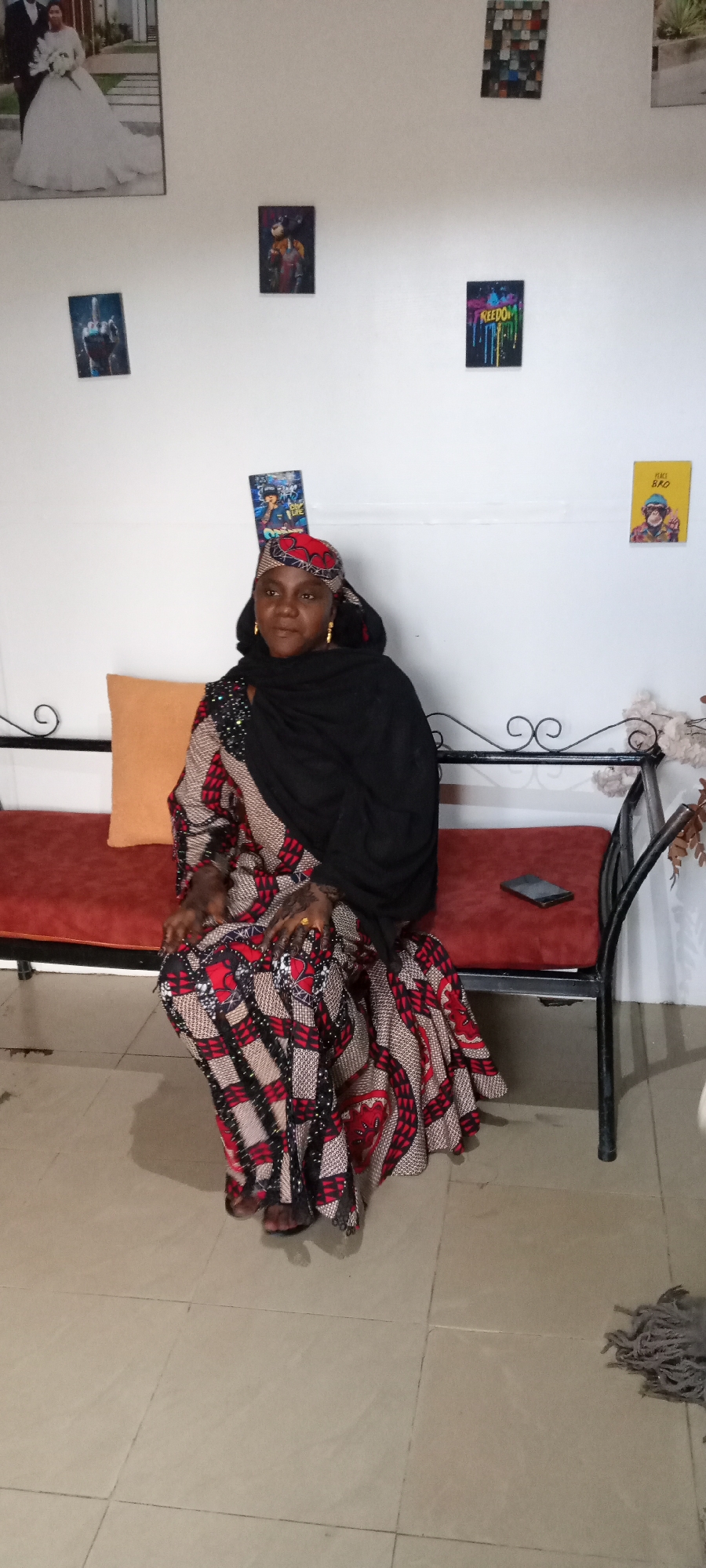 The image shows a woman seated on a red padded bench with a black metal frame. She is wearing a patterned dress with a black shawl and a headscarf. The background is a white wall with several framed pictures. On the bench, next to the woman, is a black smartphone. The location is Unknown, Unknown.