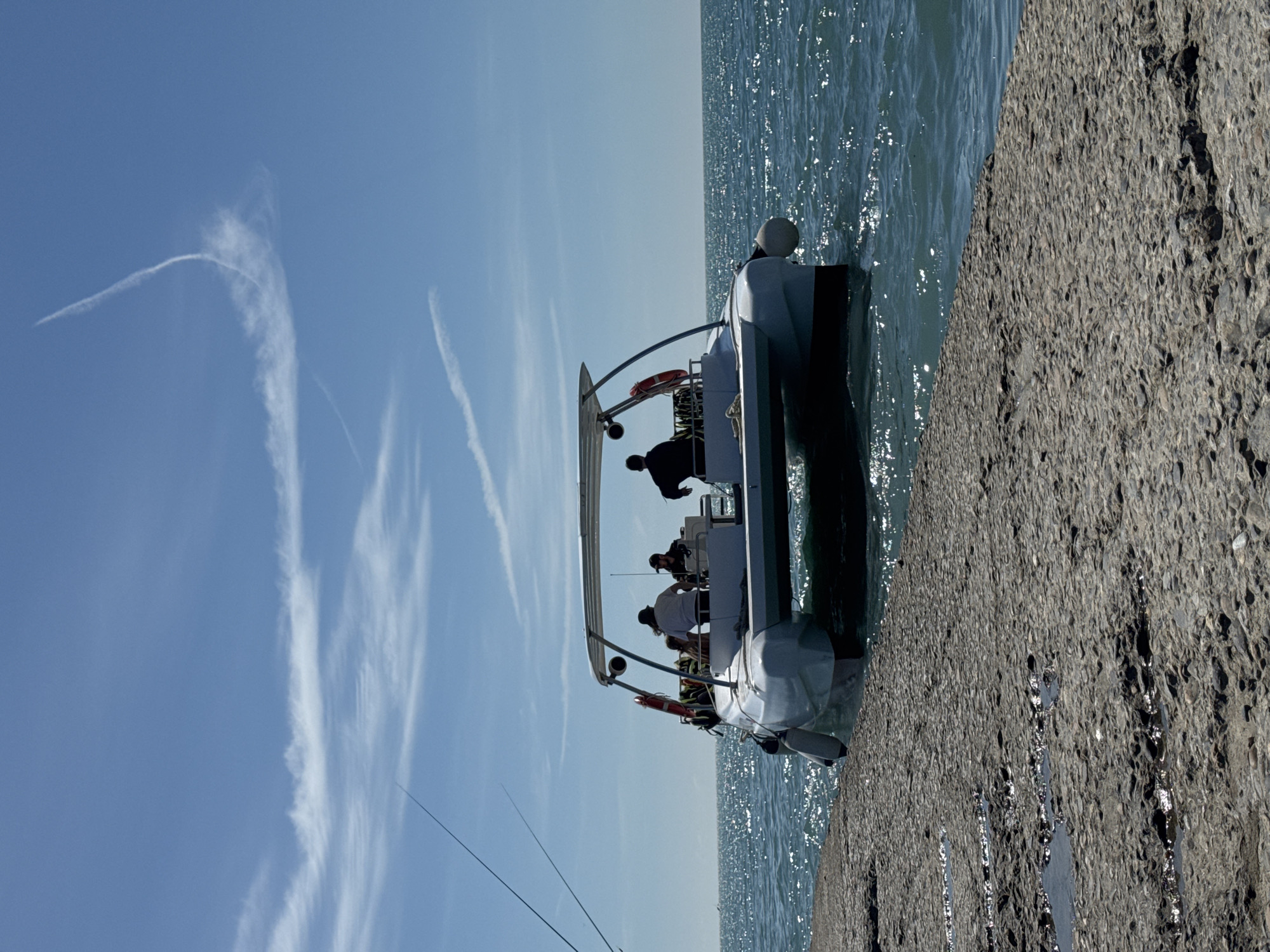 The image shows a boat docked beside a concrete pier, possibly in the city of Sochi, Russia. Several people are visible on the boat. The water is calm with sunlit ripples, and the sky is blue with contrails. Fishing rods are visible on the left. The pier is constructed of weathered concrete.