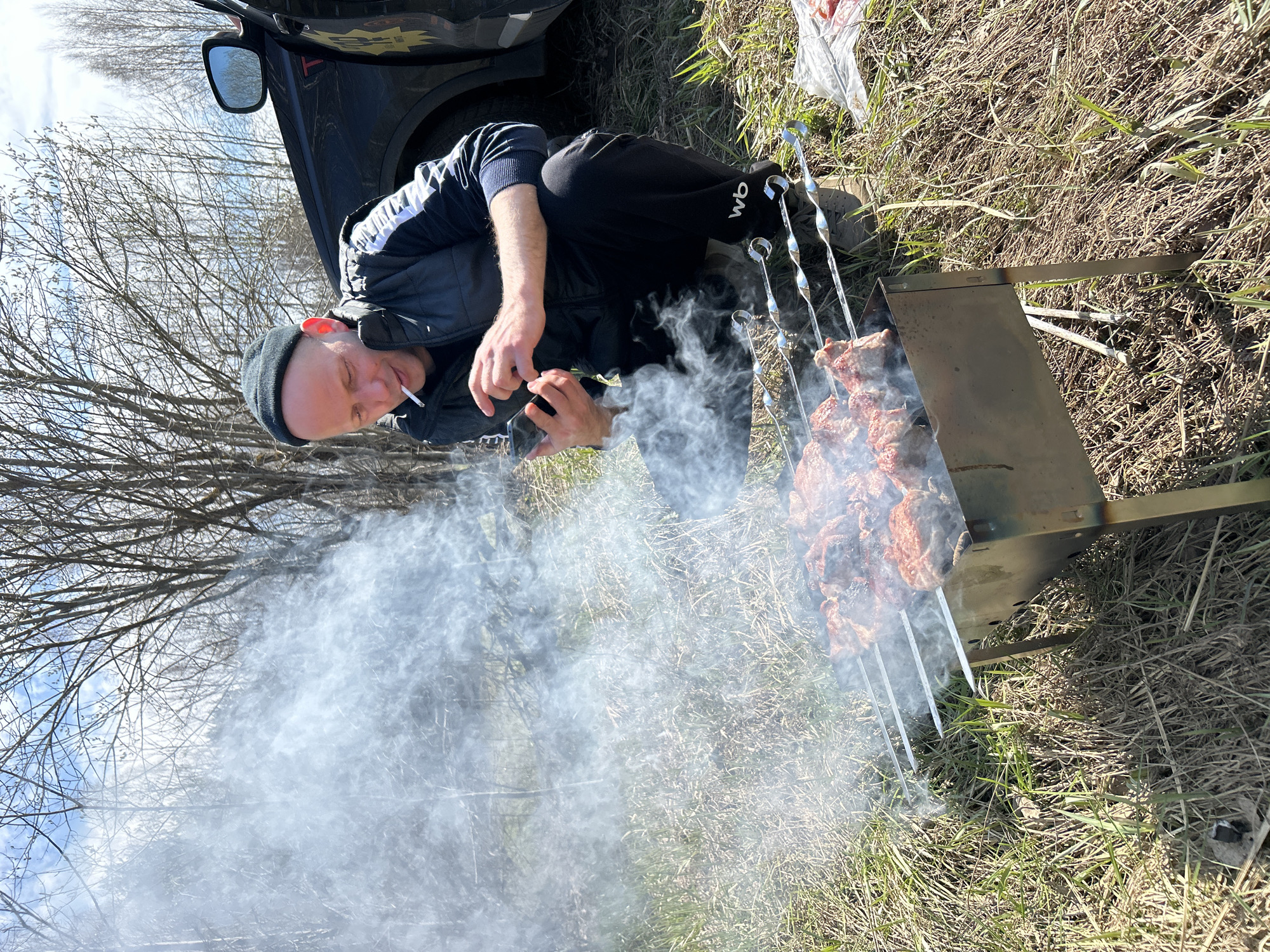 The image depicts a man in Unknown, Unknown, seated on the ground next to a barbecue grill. He is wearing a black beanie, a dark jacket with white stripes on the sleeves, and dark pants with the letters "WB" on the lower leg. He holds a cigarette in his mouth and is either lighting another cigarette or using a mobile phone with his hands. Skewers with meat are placed on the grill, producing a significant amount of smoke that obscures the foreground. A dark car is partially visible in the upper left corner of the image. The ground is covered with dry grass and some green vegetation. Bare trees are visible in the background.